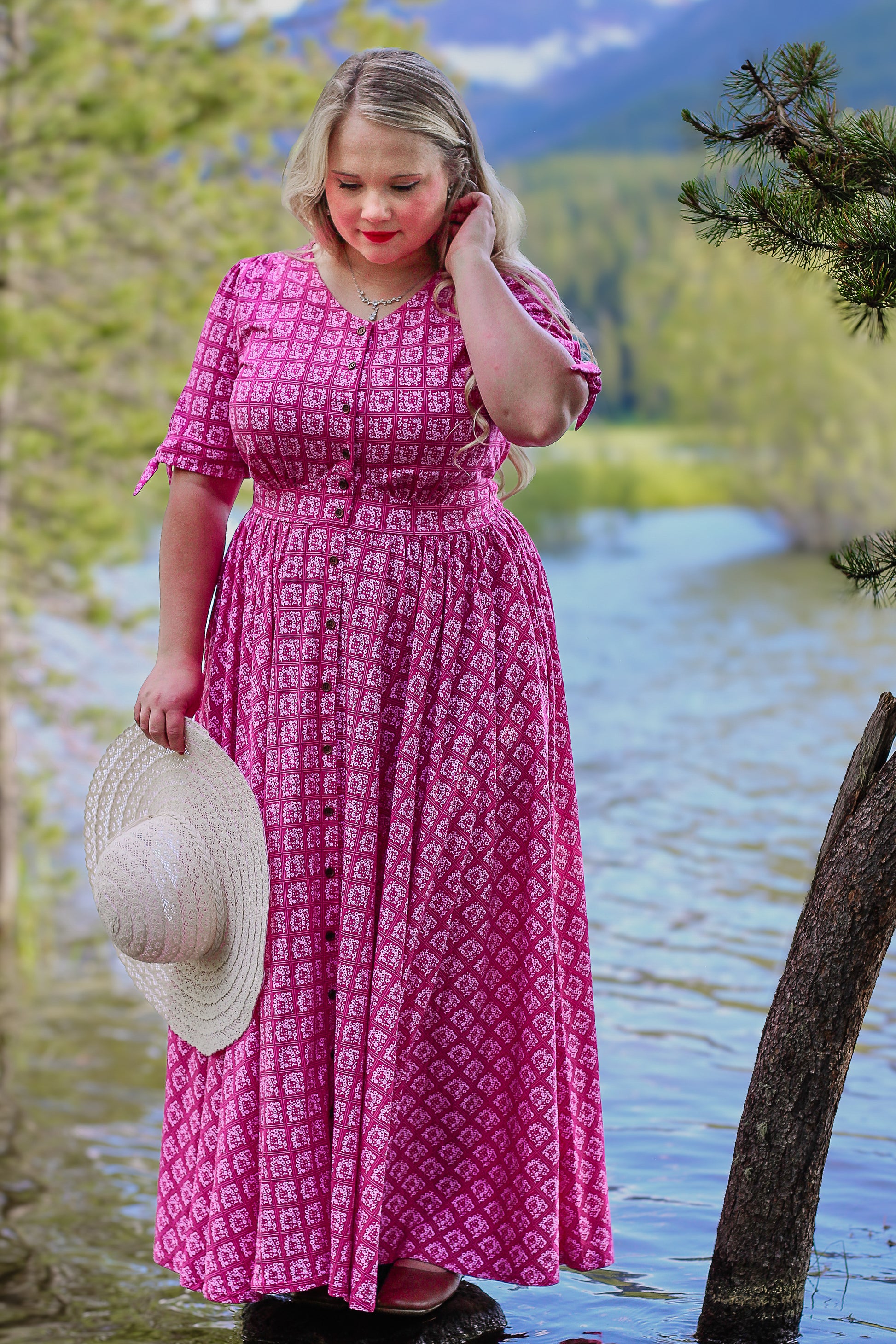 Woman in a pink modest nursing dress standing by a body of water with trees and mountains in the background