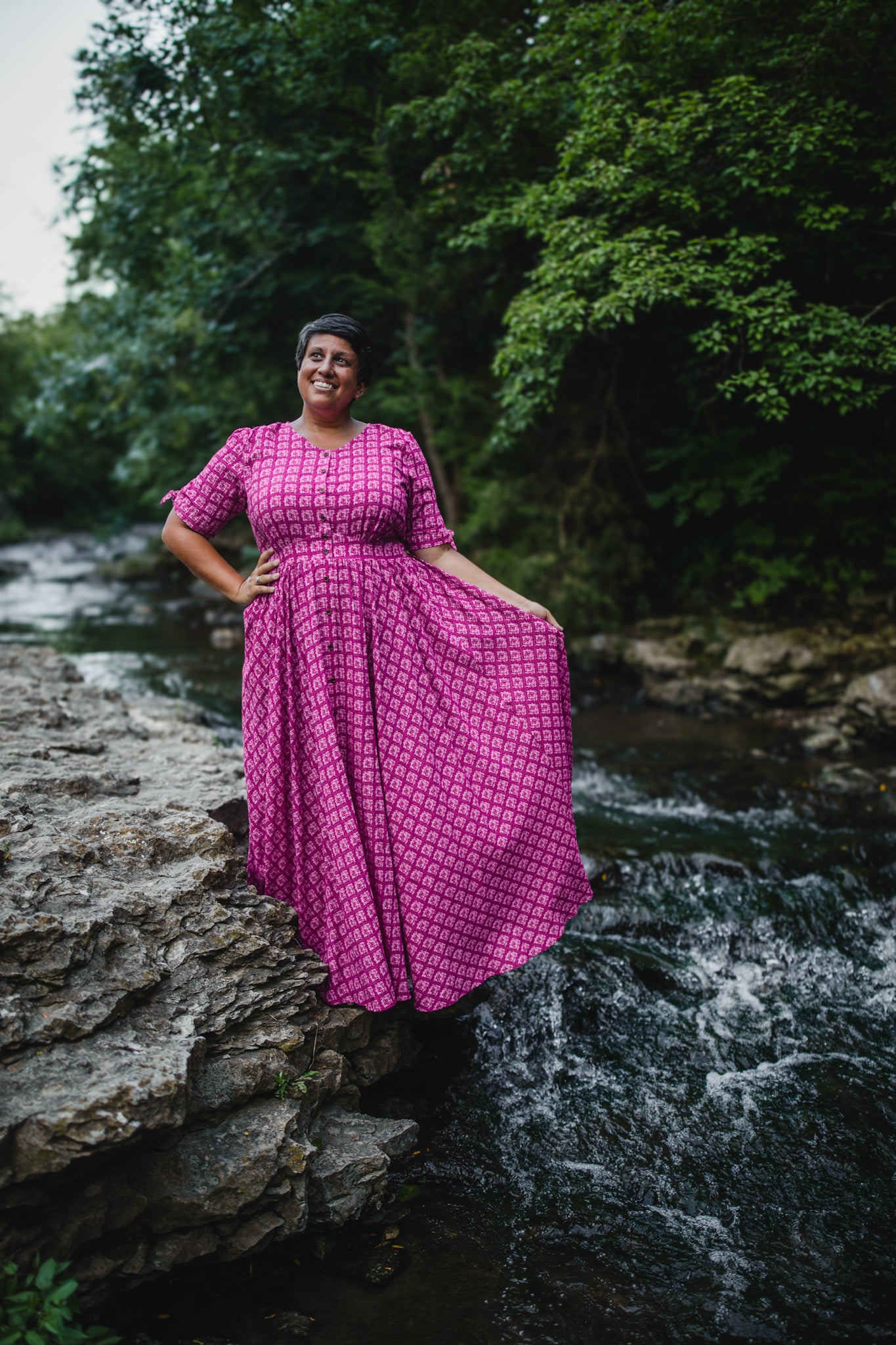 Woman in a pink modest nursing dress standing on rocks by a stream with greenery in the background