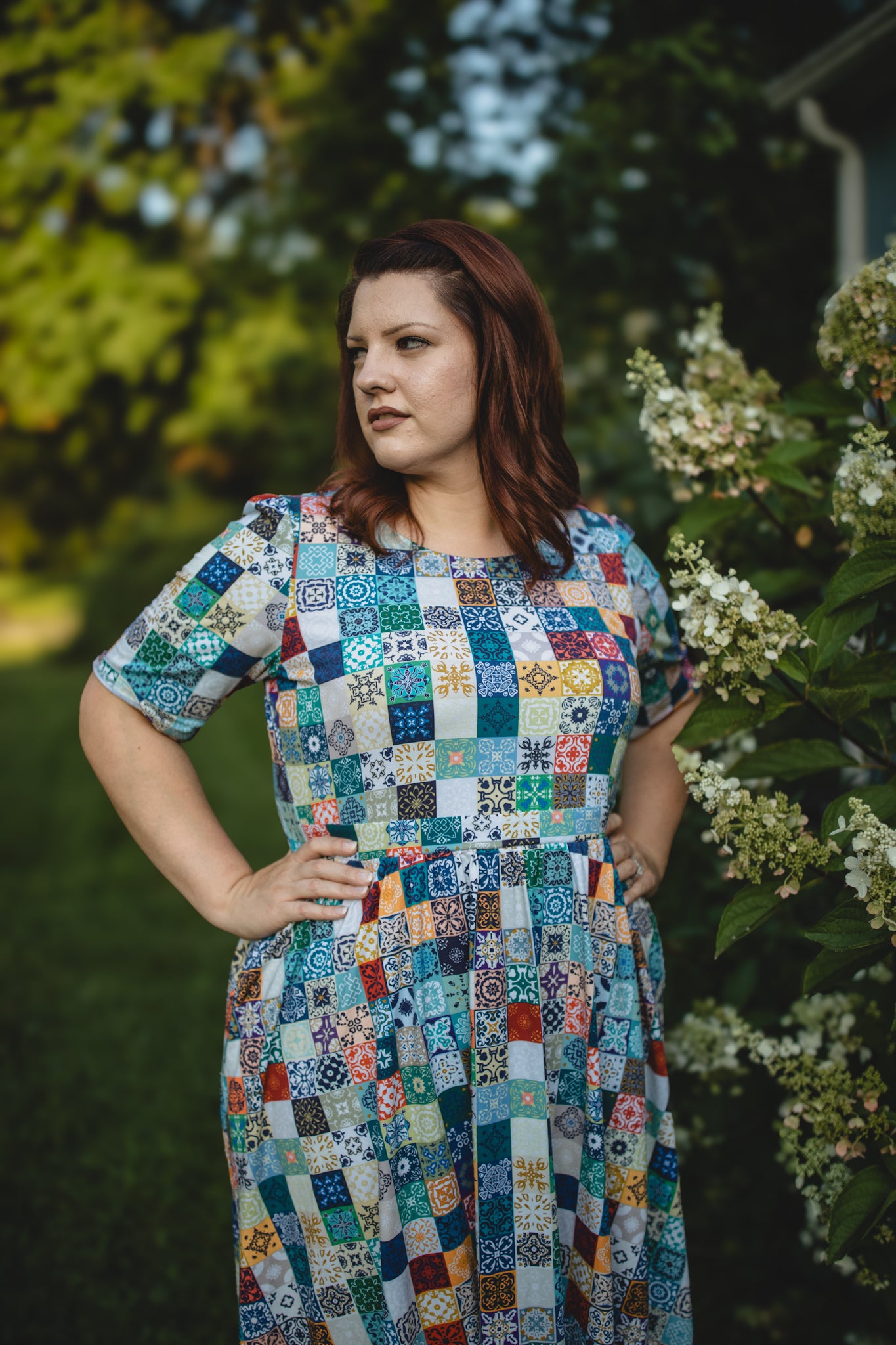 Woman wearing a colorful patchwork modest nursing dress standing outdoors with greenery and flowers in the background