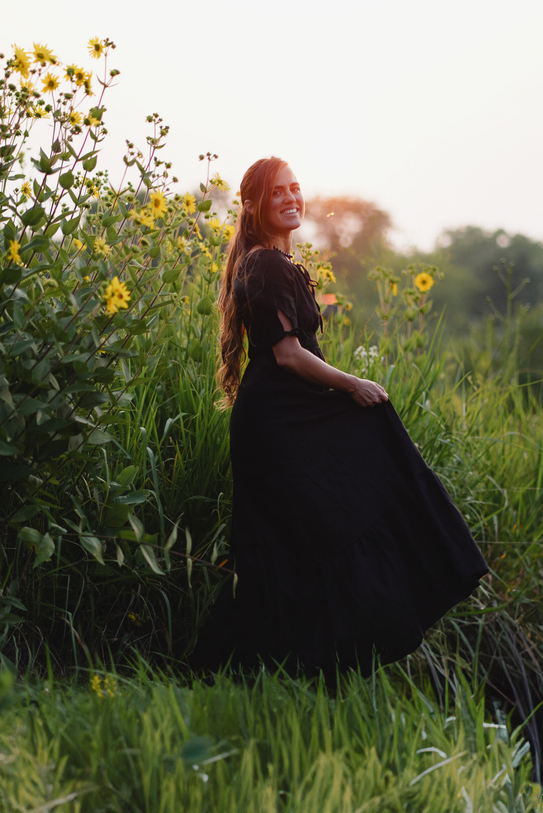 Woman in a black modest nursing dress standing in a field of tall grass and wildflowers.