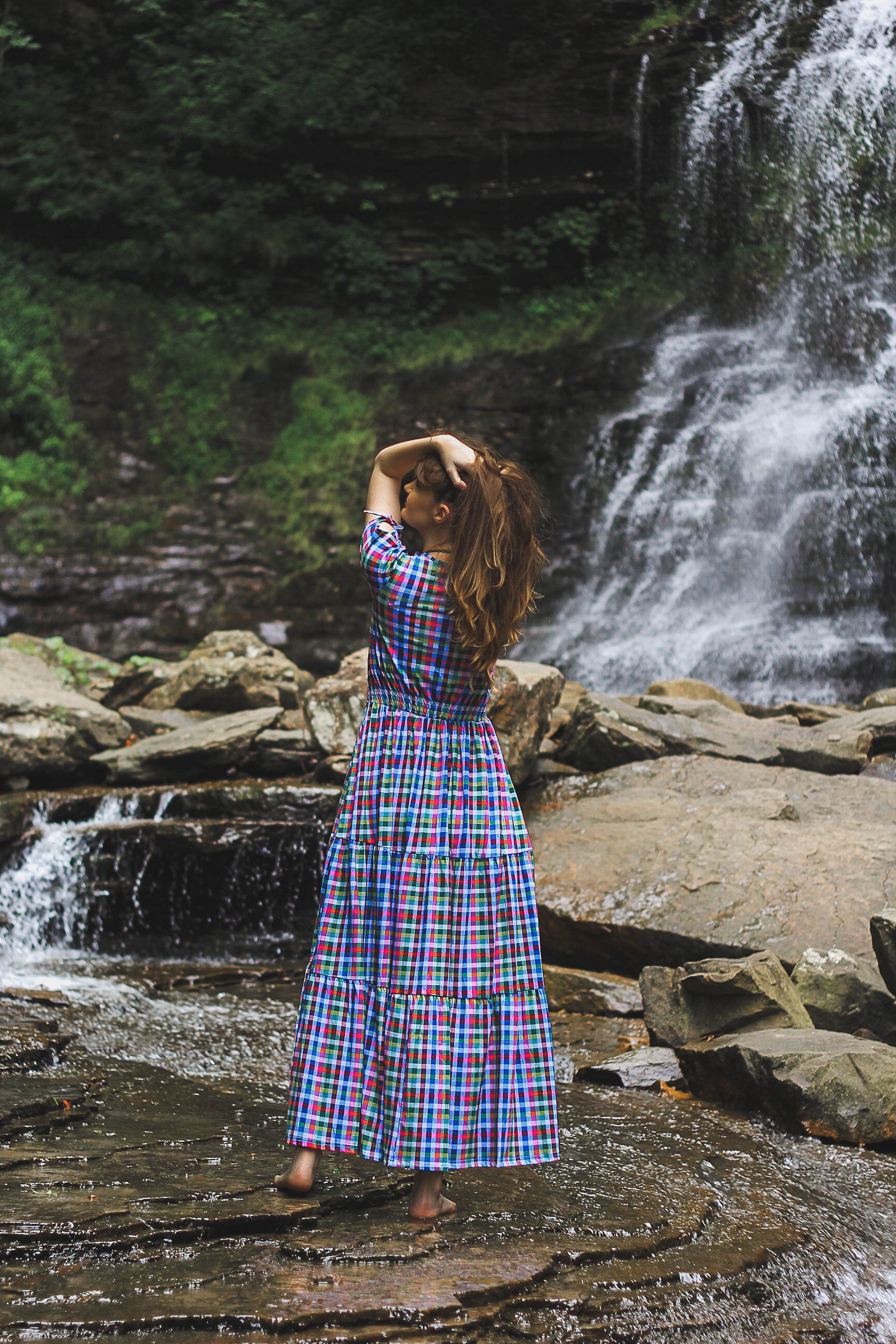Woman in a plaid modest nursing dress standing by a waterfall in a natural setting