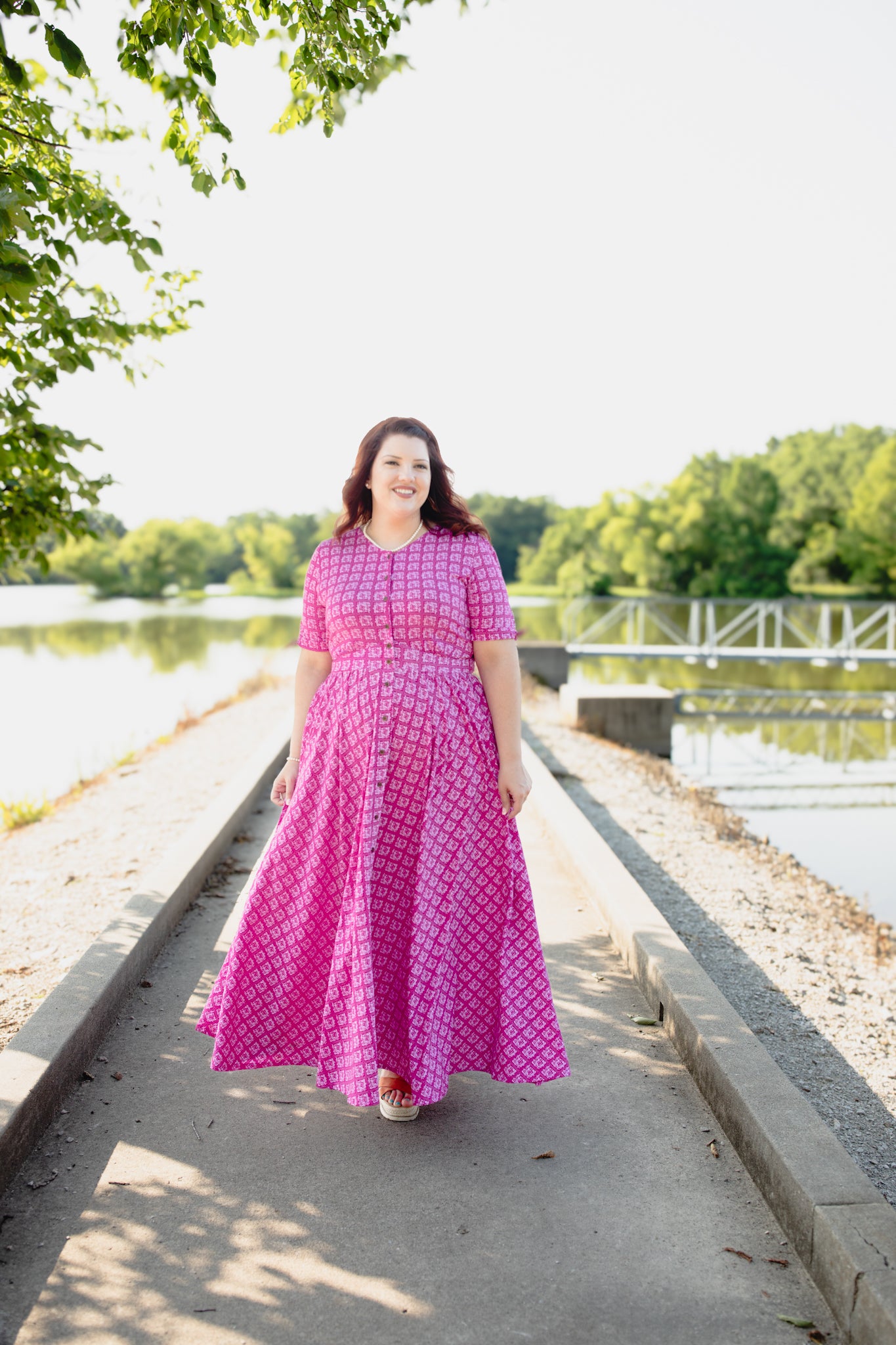Woman in a pink modest nursing dress standing by a lake with trees in the background