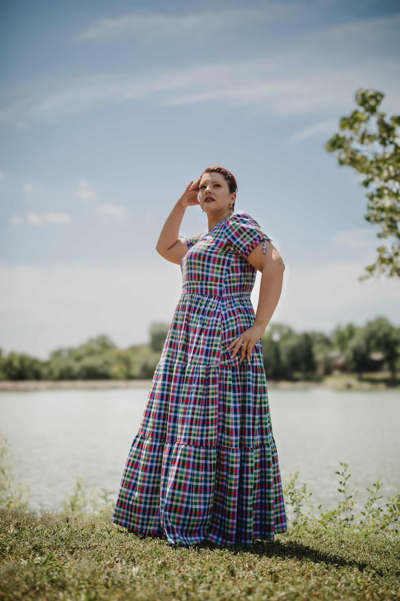 Woman in a plaid modest nursing dress standing outdoors by a lake with trees in the background