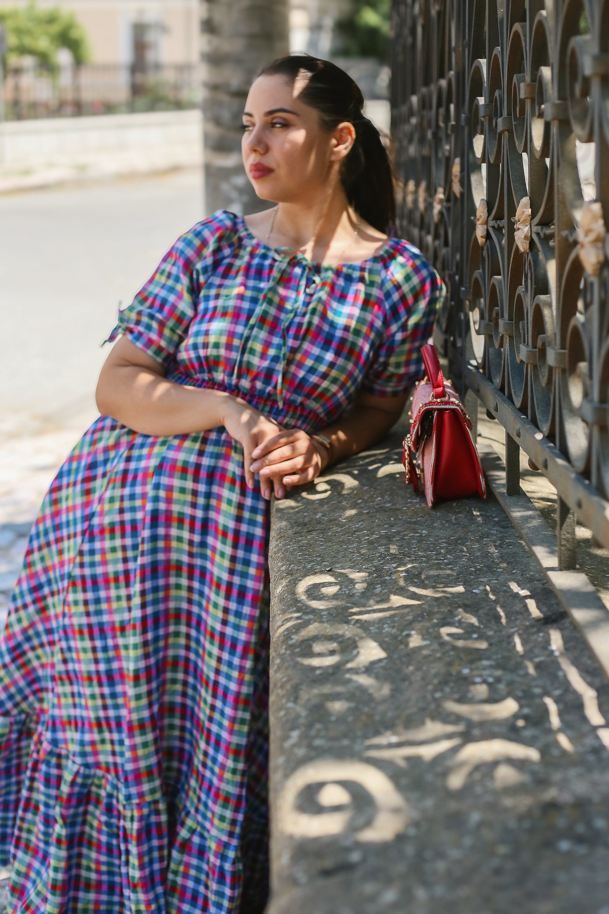 Woman in a colorful modest nursing dress leaning against a metal fence on a street.