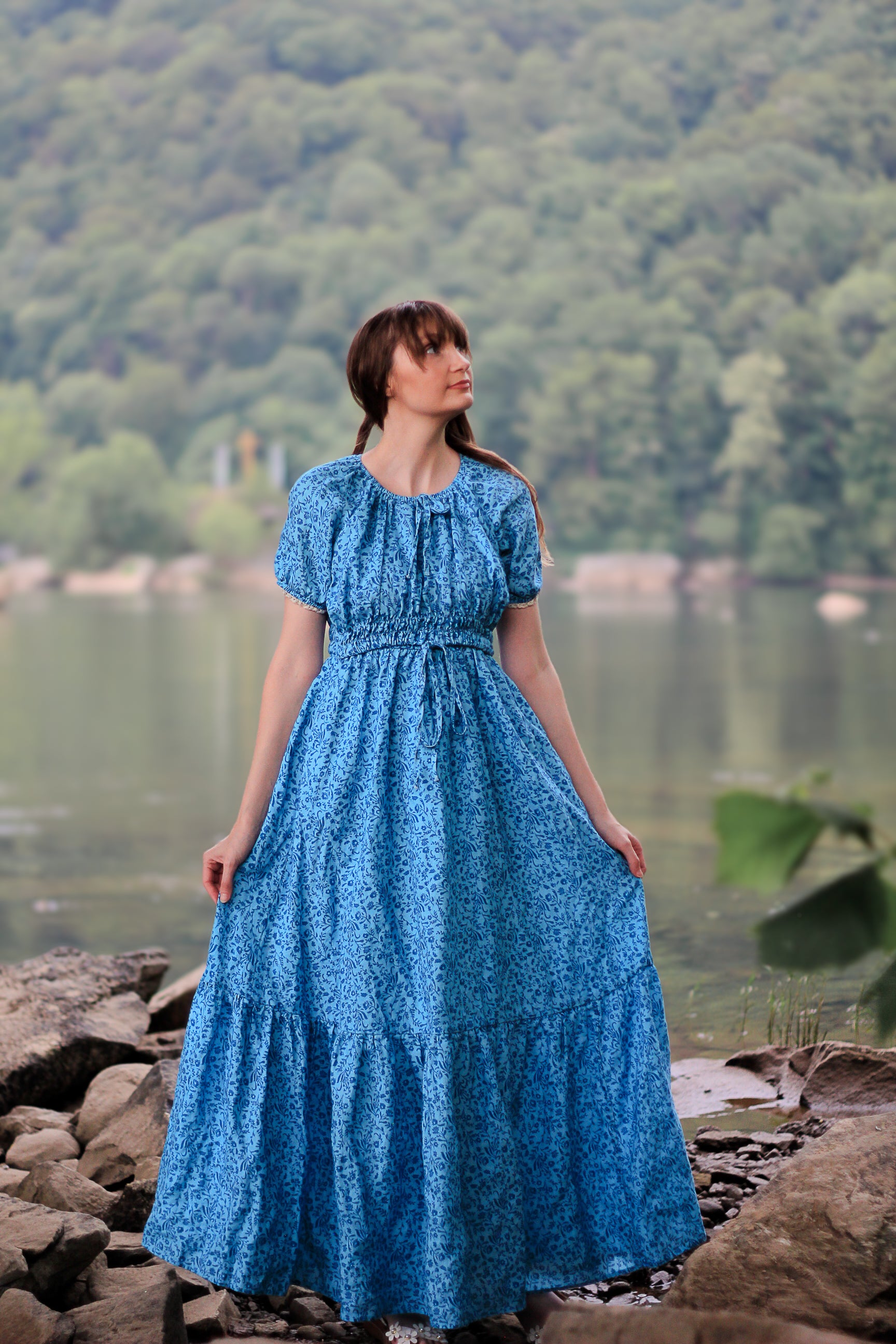 Woman in a blue modest nursing dress standing by a lake with trees in the background