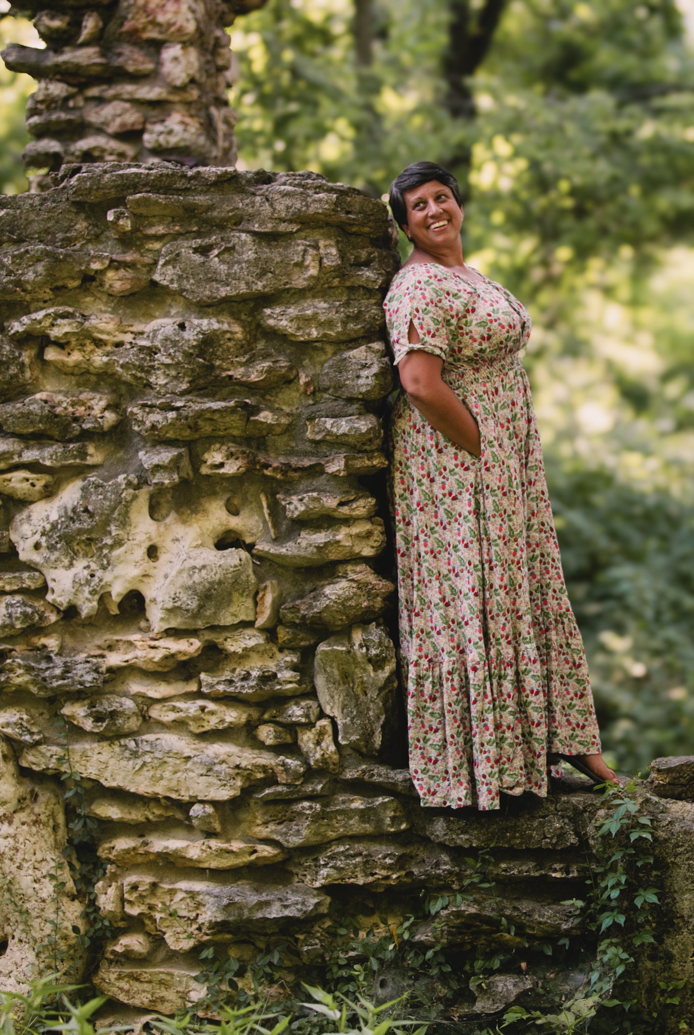 Woman in a floral modest nursing dress leaning against a stone wall in a forest setting