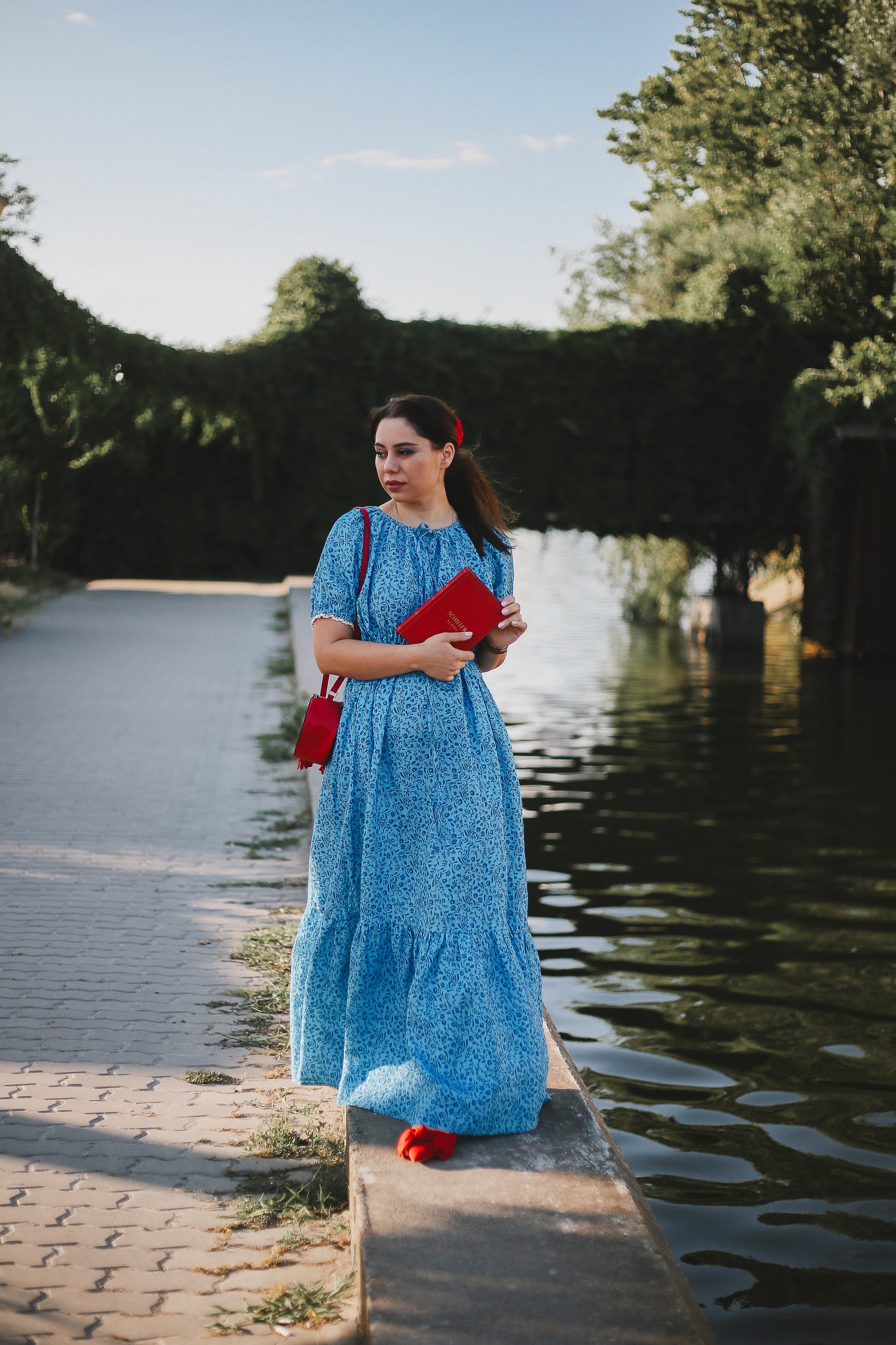 Woman in a blue modest nursing dress standing by a body of water with trees in the background