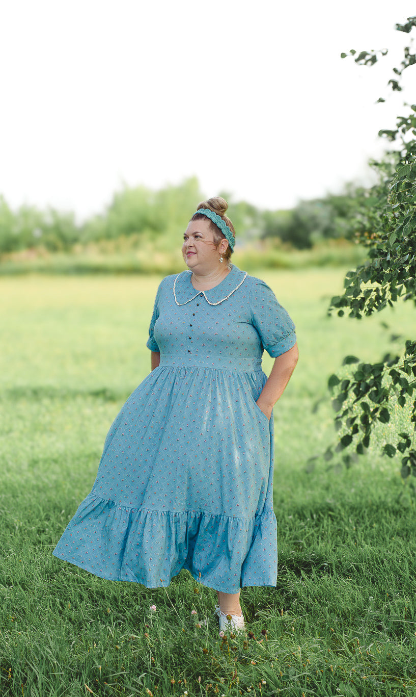 Woman in a blue modest nursing dress standing in a grassy field with trees in the background