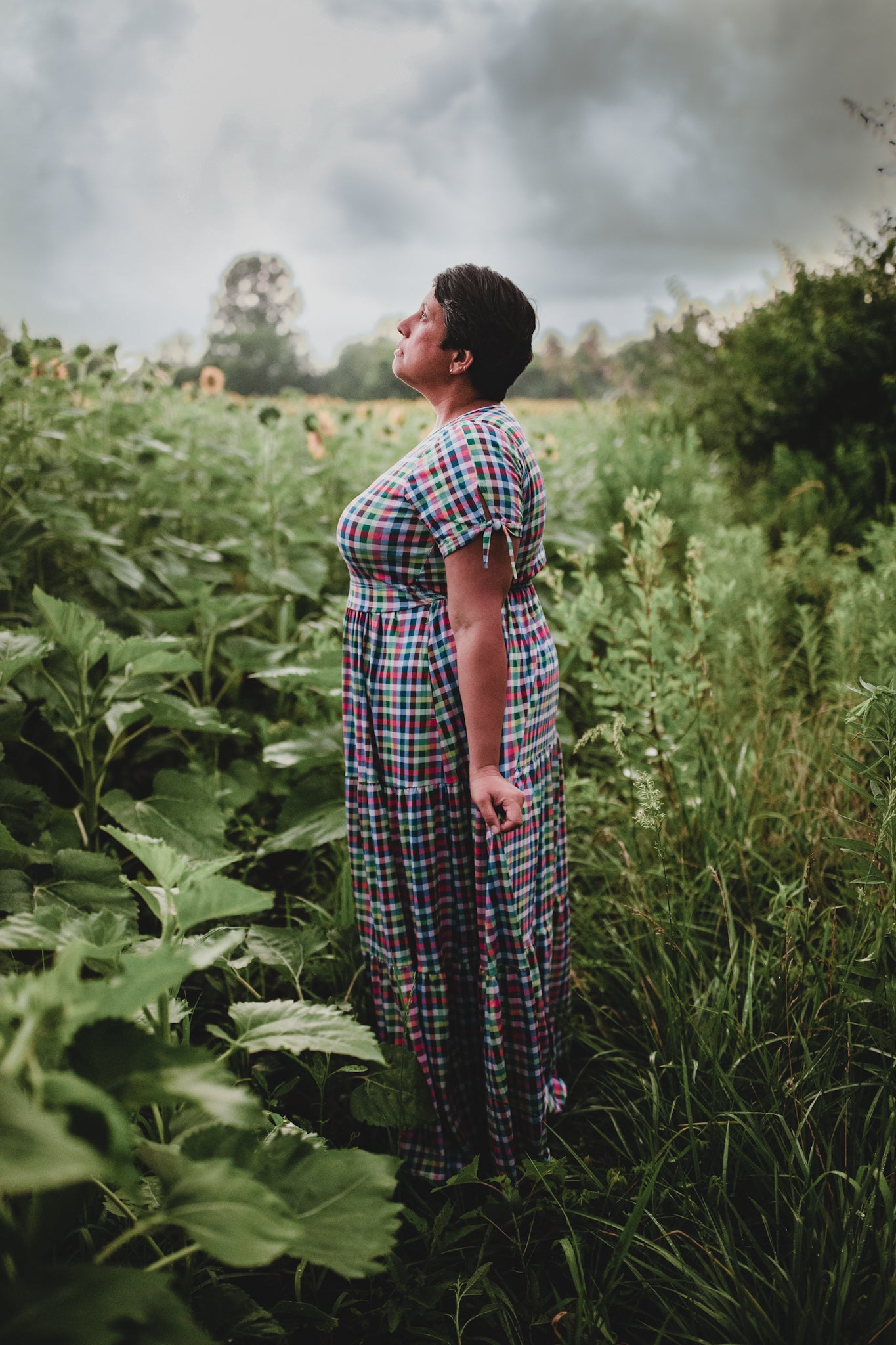Woman in a plaid modest dress standing in a field of tall green plants with a cloudy sky.