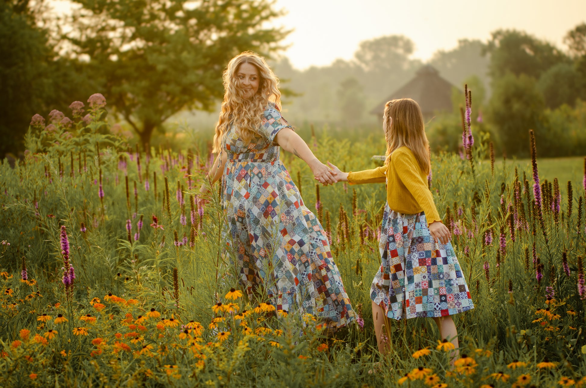 Mother and daughter holding hands in a field of flowers during sunset wearing modest dresses.
