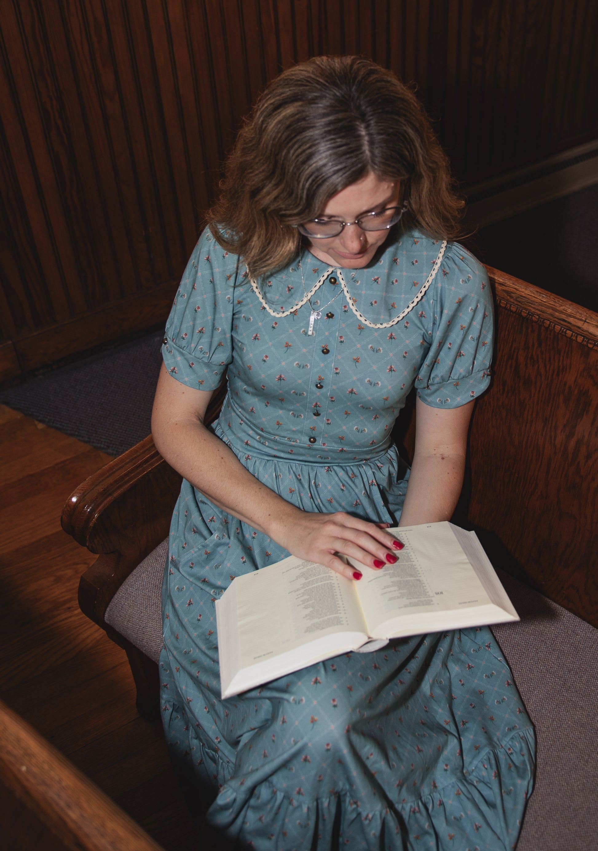 Woman in a blue modest nursing dress reading a book in a wooden pew.