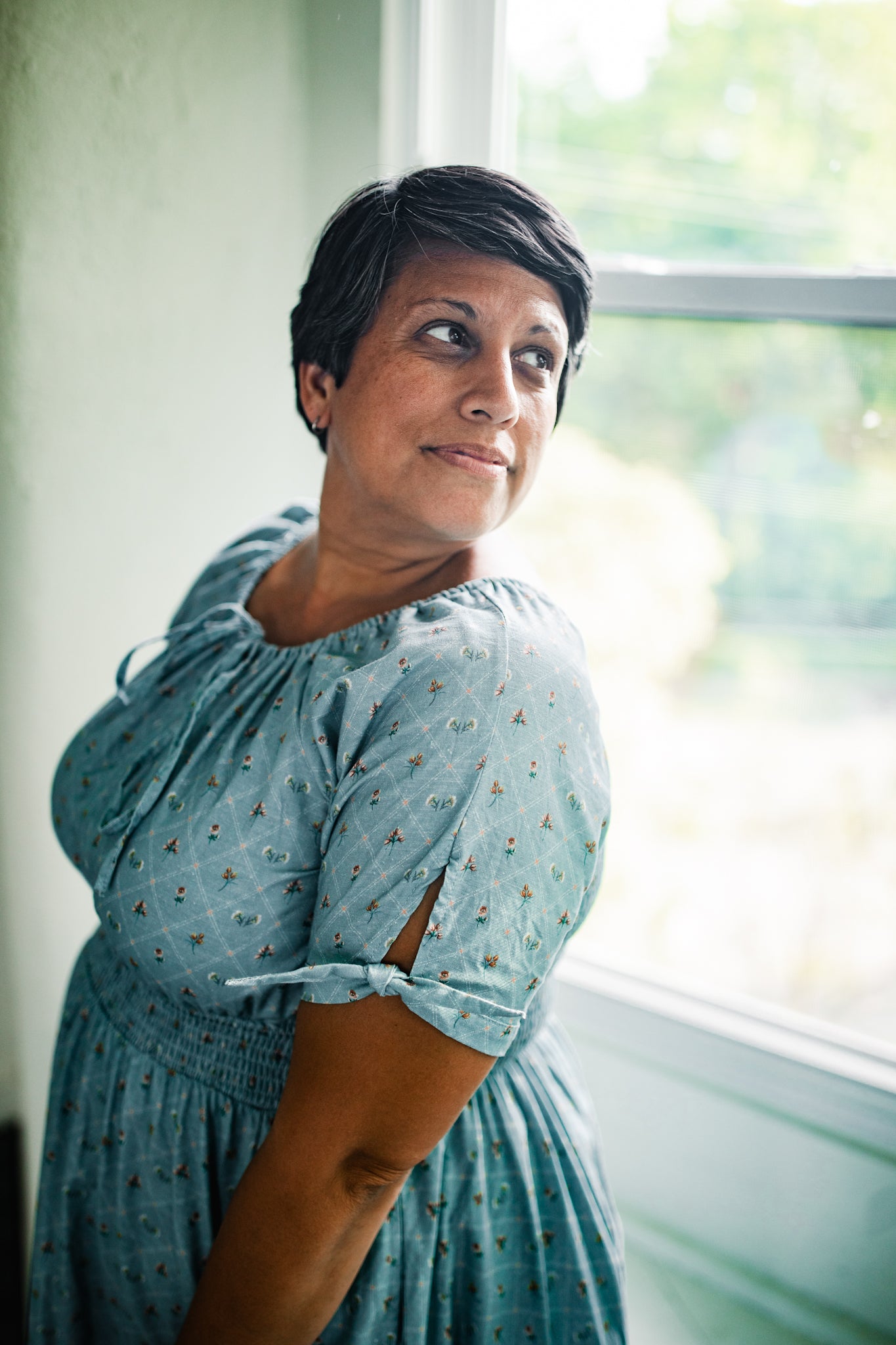 Woman in a blue modest nursing dress standing by a window with a blurred outdoor background