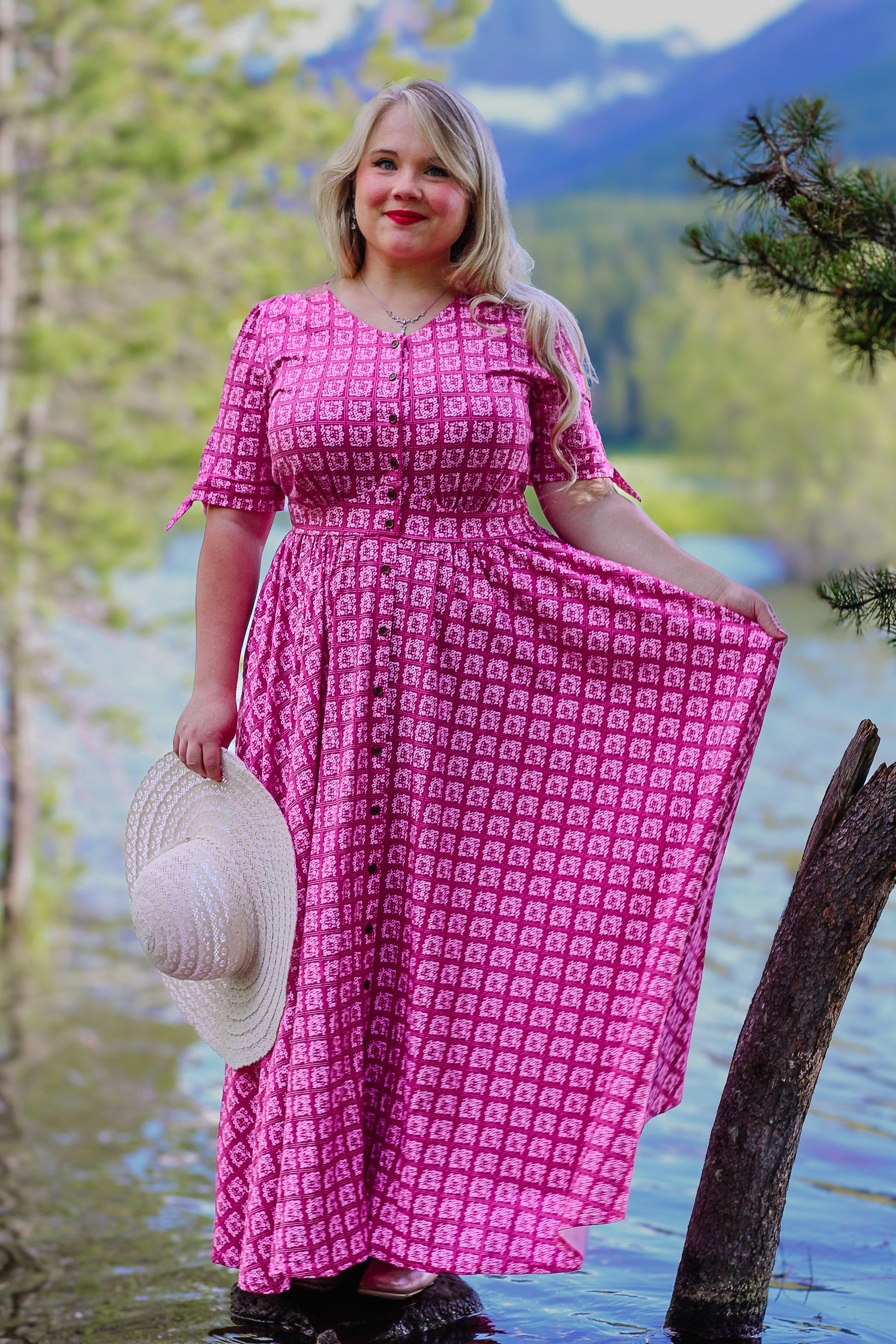 Woman in a pink modest nursing dress standing by a body of water with mountains in the background