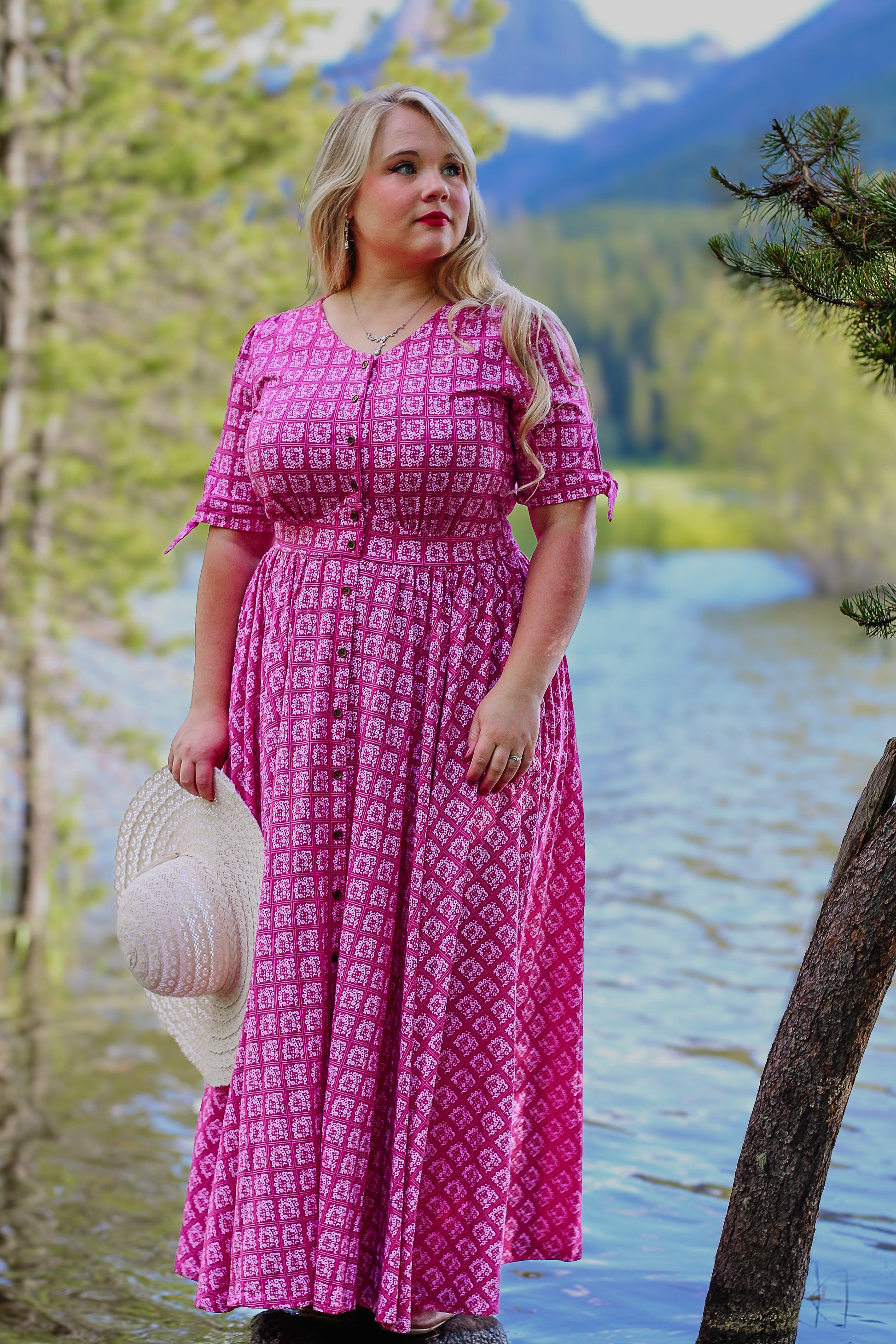 Woman in a pink modest nursing dress standing by a lake with mountains in the background