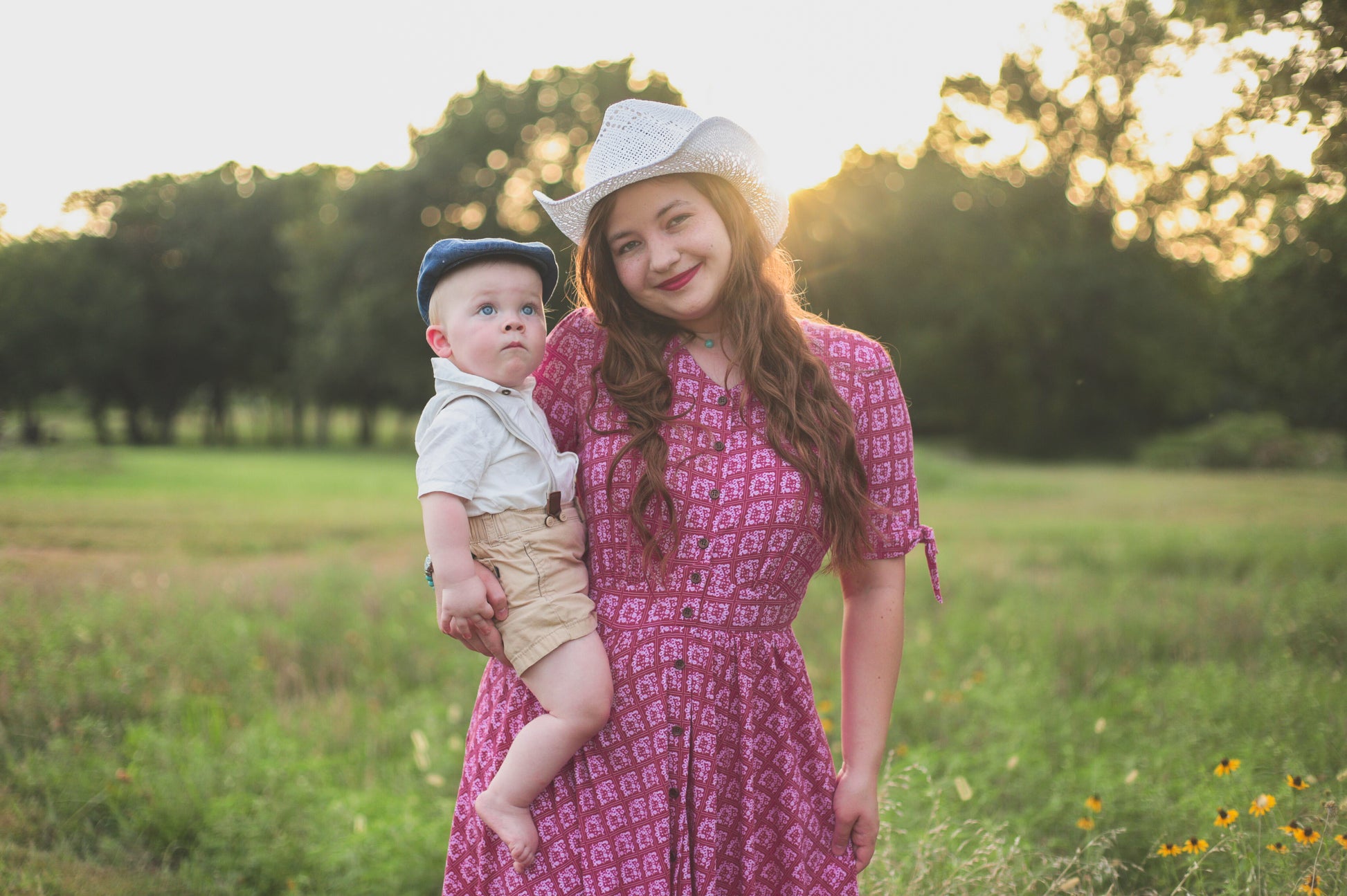 Woman in a pink modest nursing dress holding a baby in a field with trees in the background