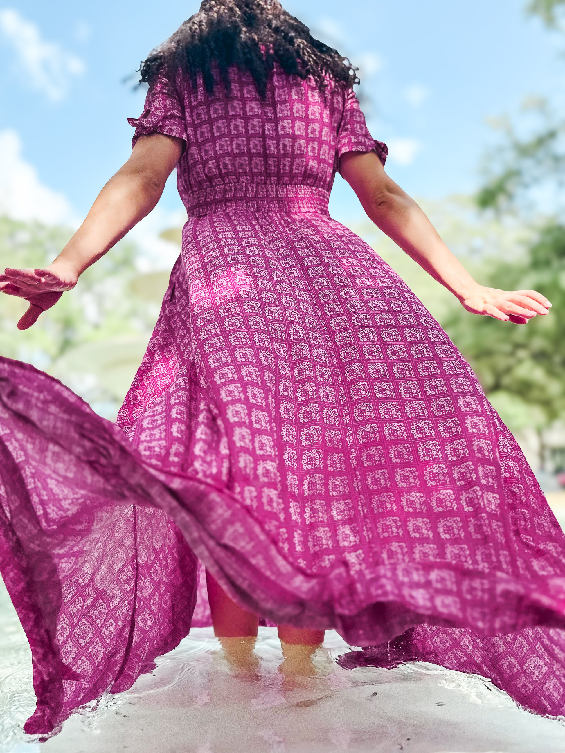 Person wearing a pink patterned modest nursing dress with arms outstretched, standing on a white surface with a blurred natural background.