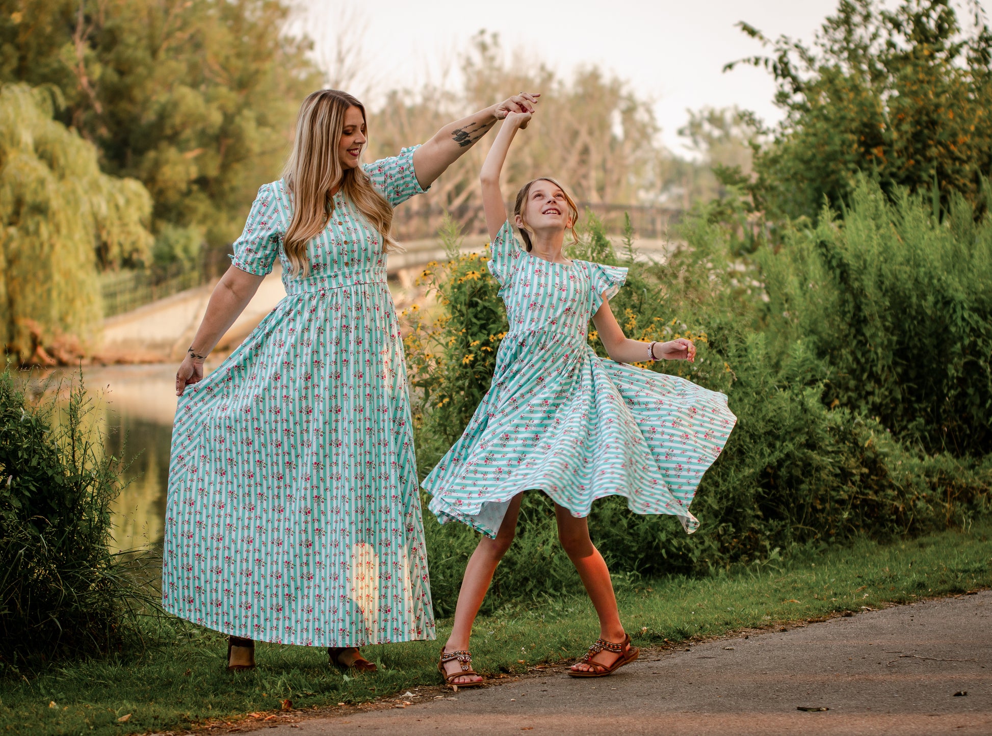 Mother and daughter in matching modest dresses dancing outdoors near a lake.