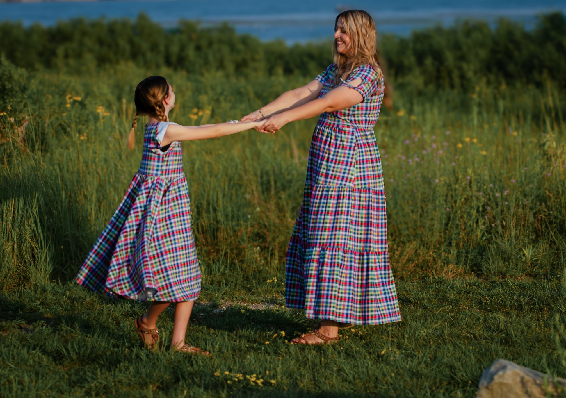 Mother and daughter in plaid modest dresses holding hands in a grassy field.