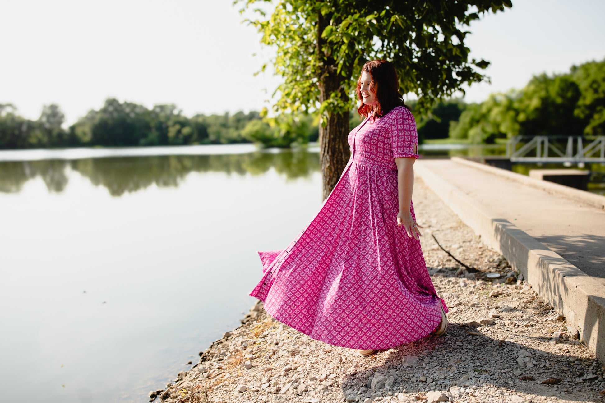 Woman in a pink modest nursing dress standing by a lake with trees in the background