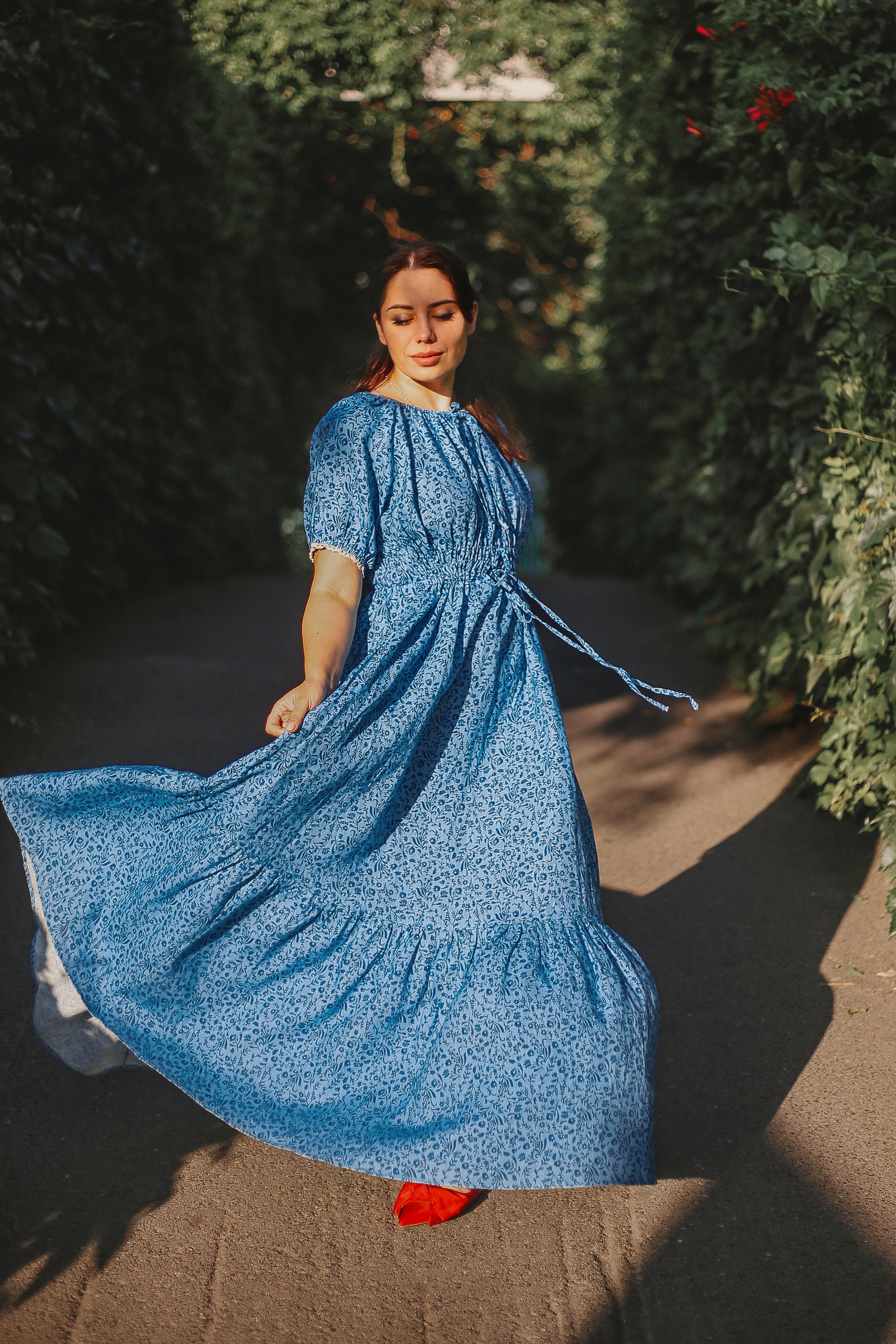 Woman in a blue modest nursing dress standing outdoors with greenery in the background