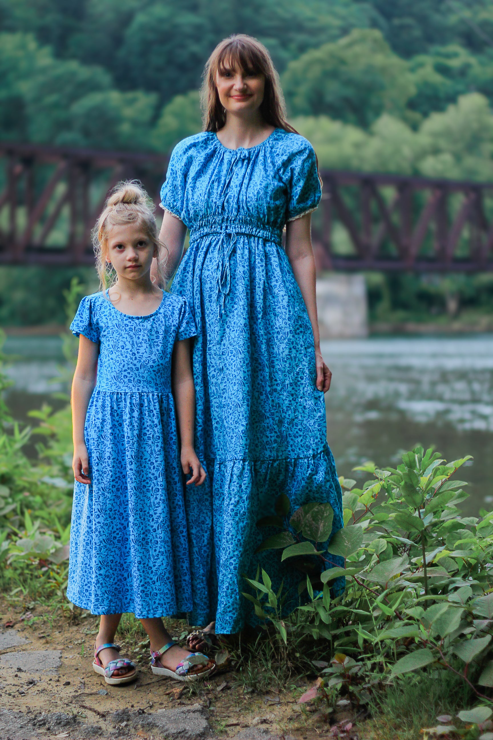 Woman and young girl in matching blue modest dresses standing by a lake with a bridge in the background.