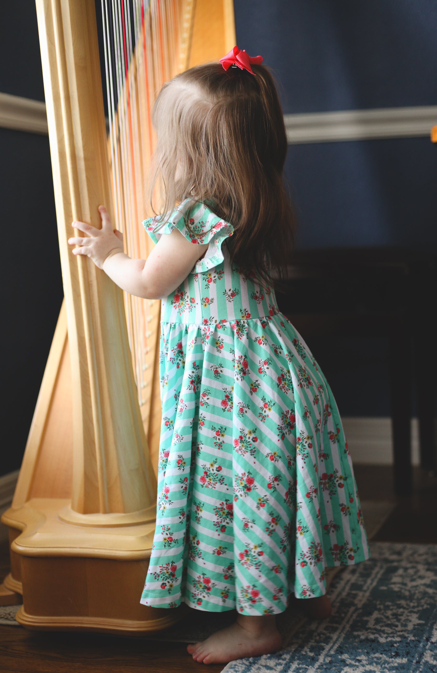 Child in a floral modest dress standing next to a harp in a room with a blue wall.