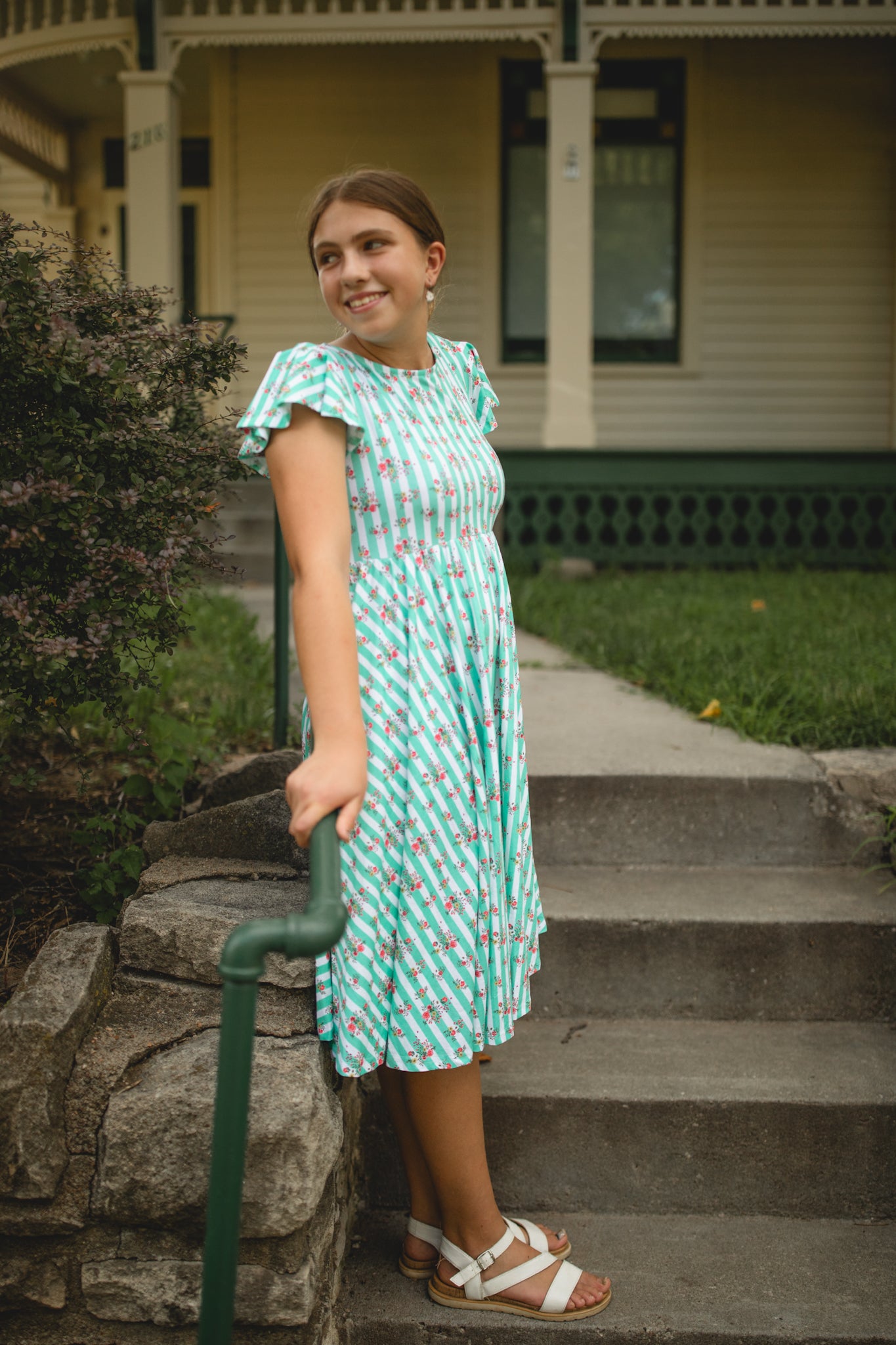 Woman in a green patterned modest dress standing on steps outside a house.