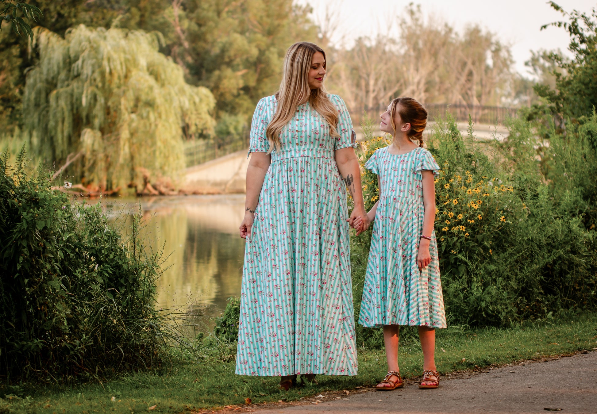 Woman and child holding hands in matching modest dresses by a pond