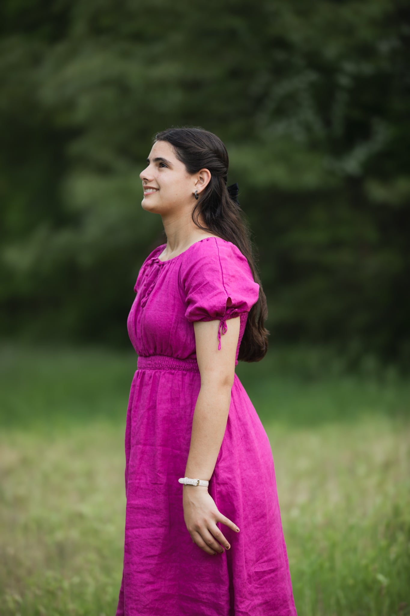 Woman in a pink modest nursing dress standing in a grassy field with blurred greenery in the background