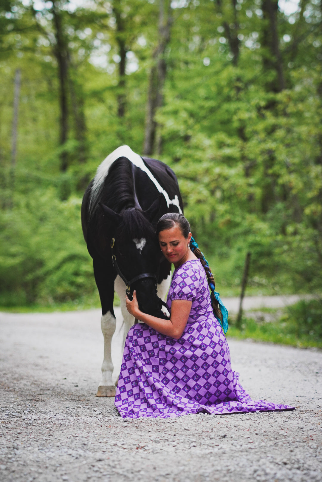 Woman in a purple modest nursing dress hugging a horse on a road surrounded by trees.