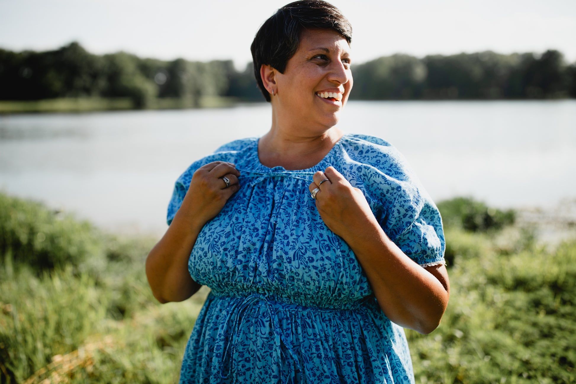 Woman in a blue modest nursing dress standing by a lake with trees in the background