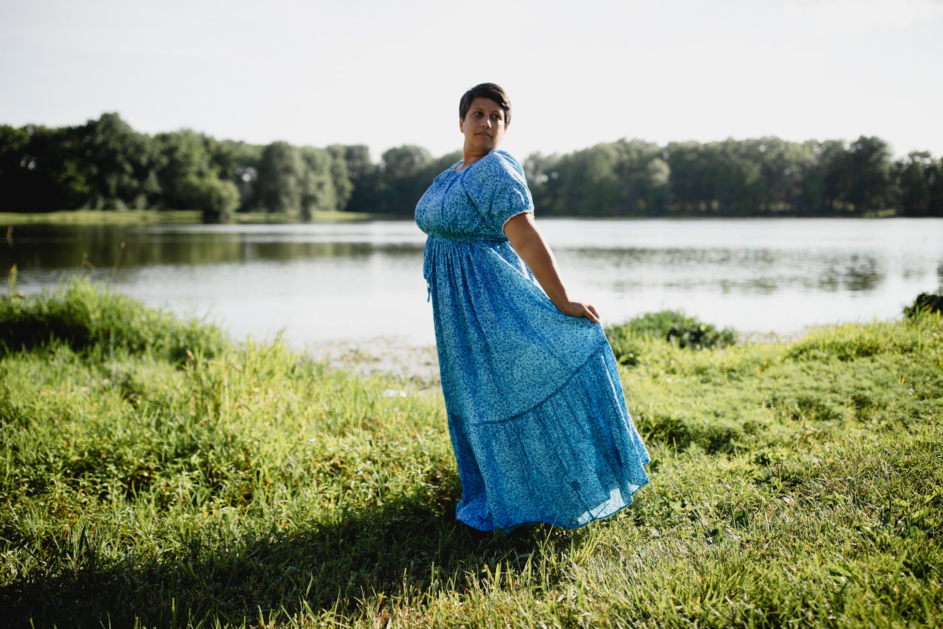 Woman in a blue modest nursing dress standing by a lake with trees in the background