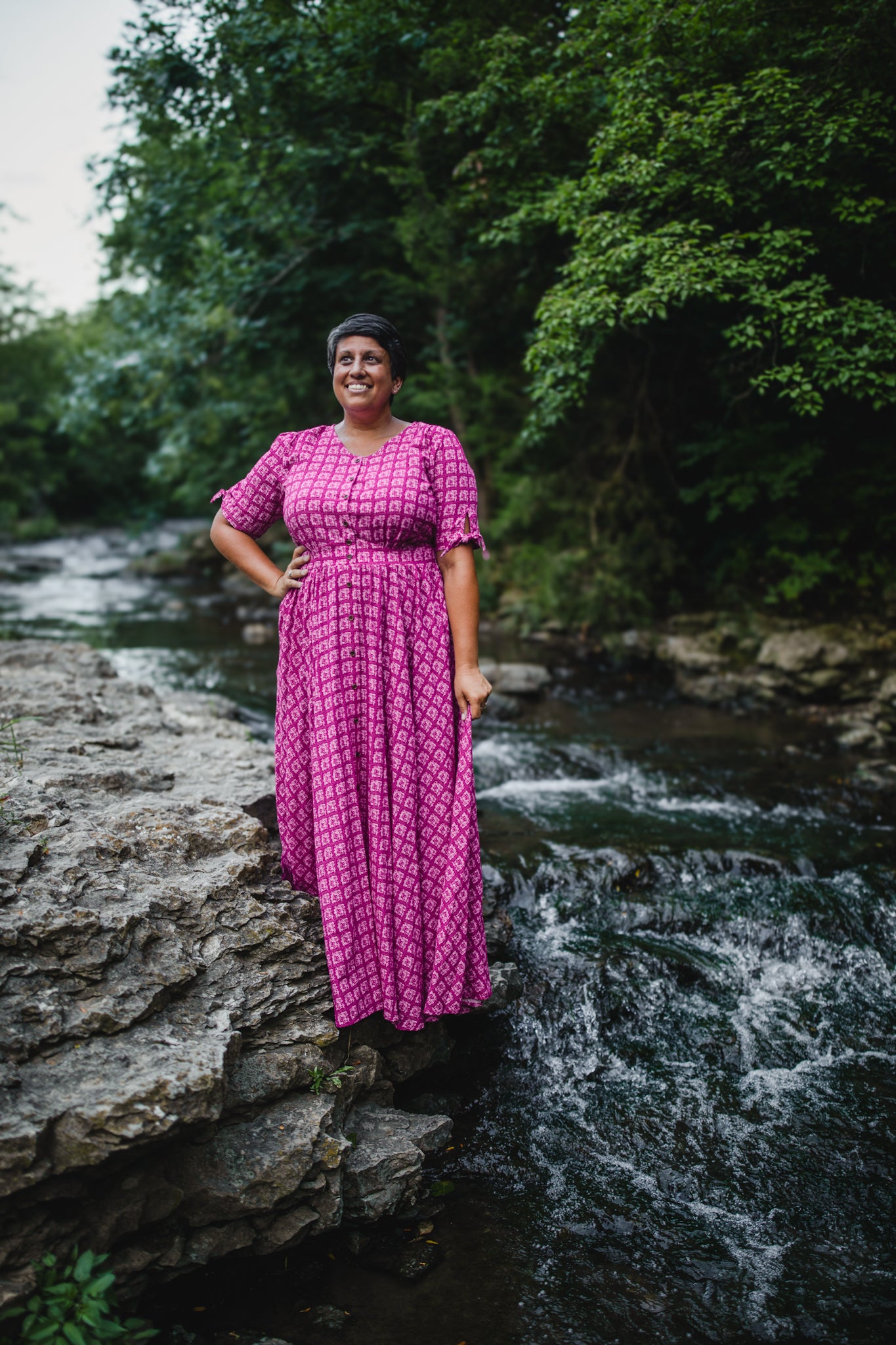 Woman in a pink modest nursing dress standing on rocks by a stream with trees in the background