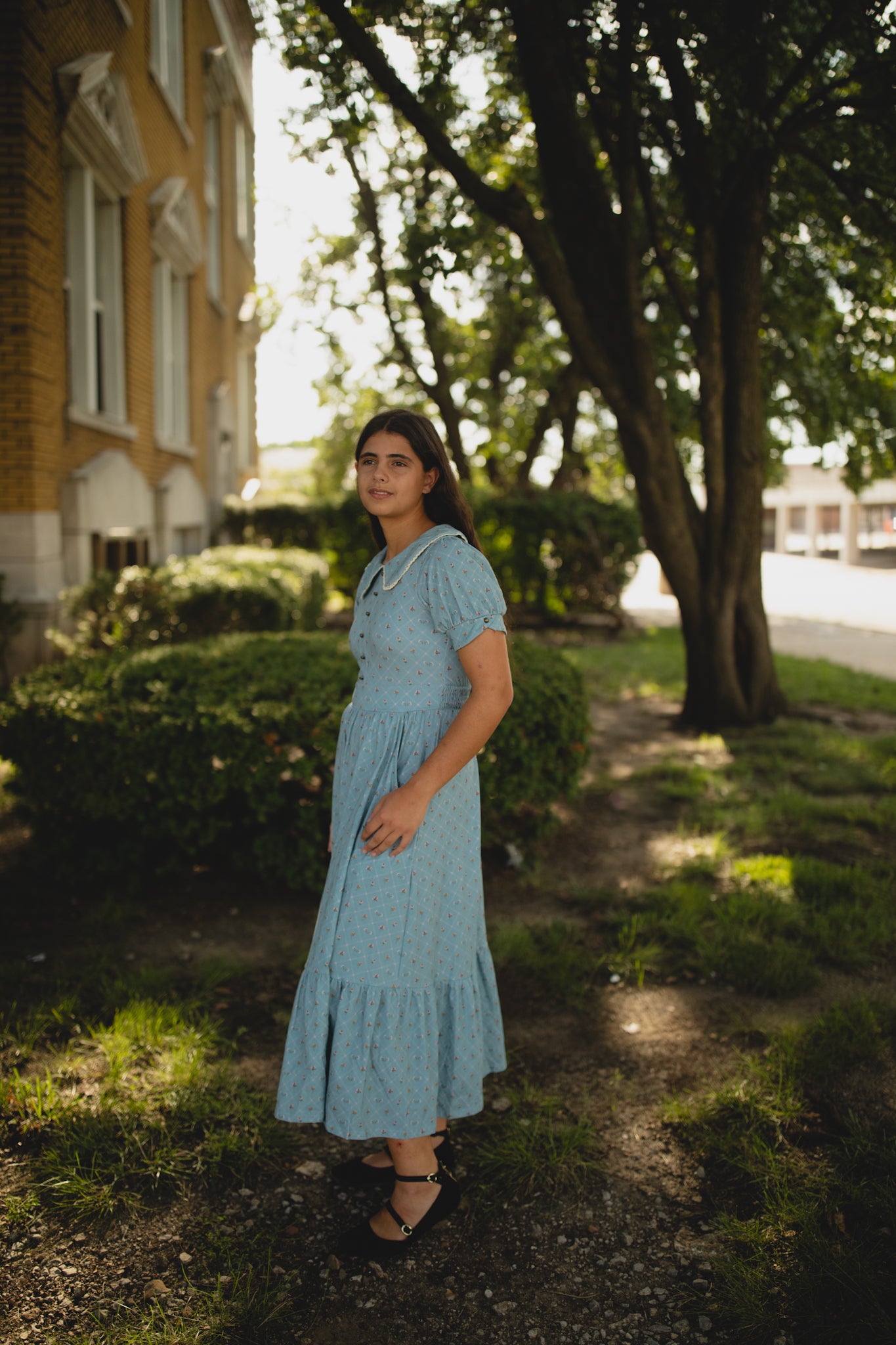 Woman in a blue modest nursing dress standing outdoors near trees and a building.
