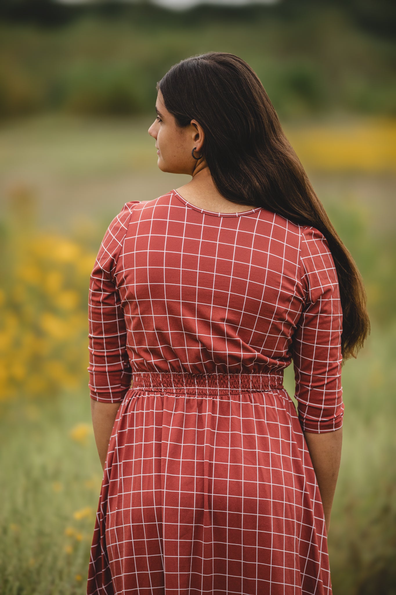 Woman wearing modest copper windowpane plaid maxi dress with 3/4 sleeves and v-neckline, standing gracefully in field setting with soft natural lighting, showcasing the nursing-friendly design and comfortable cotton blend fabric perfect for outdoor photography