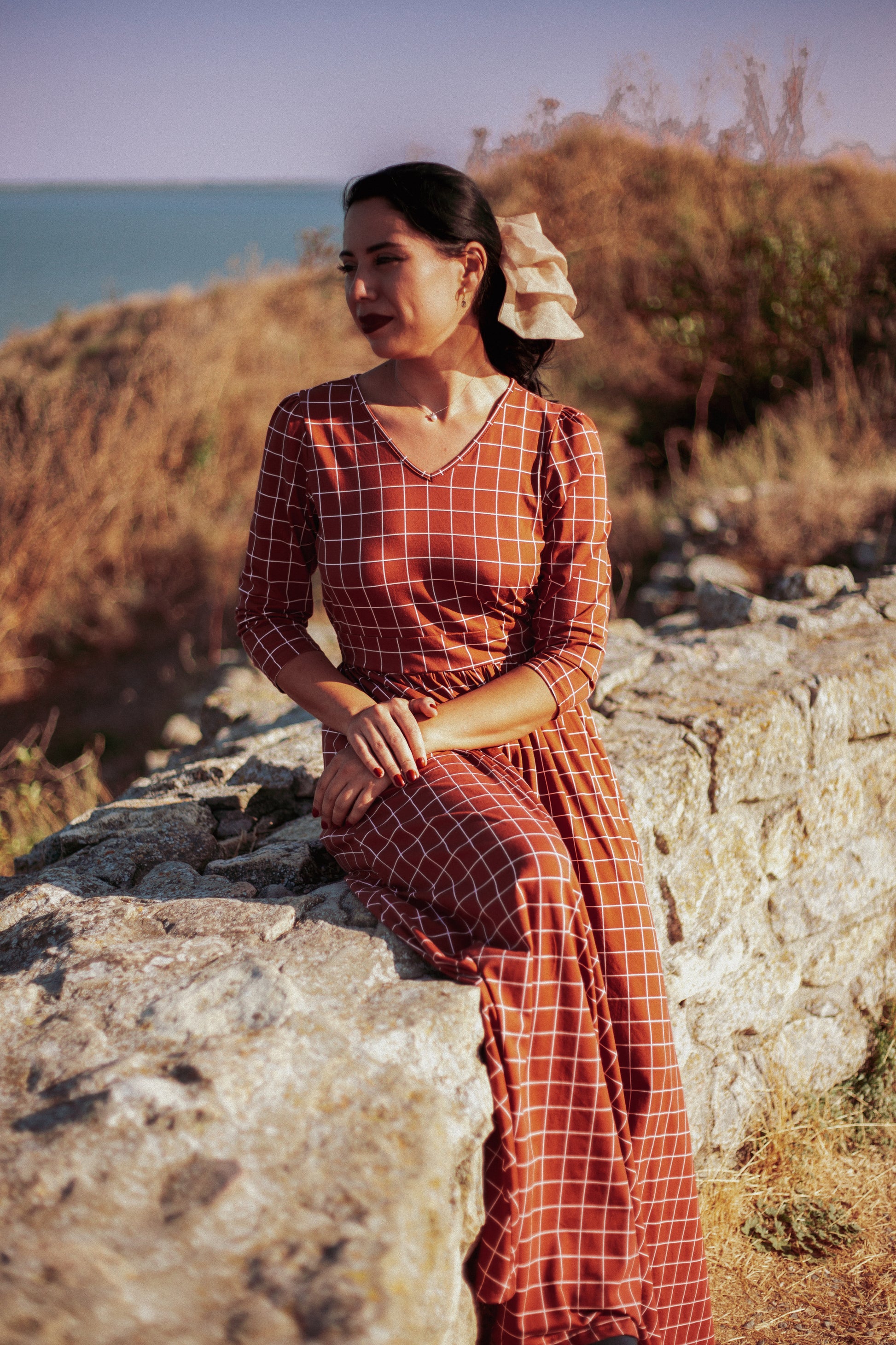 Woman wearing modest copper windowpane plaid maxi dress with 3/4 sleeves and v-neckline, sitting peacefully on coastal rocks by the sea, showcasing the nursing-friendly design and comfortable stretch fabric perfect for outdoor activities
