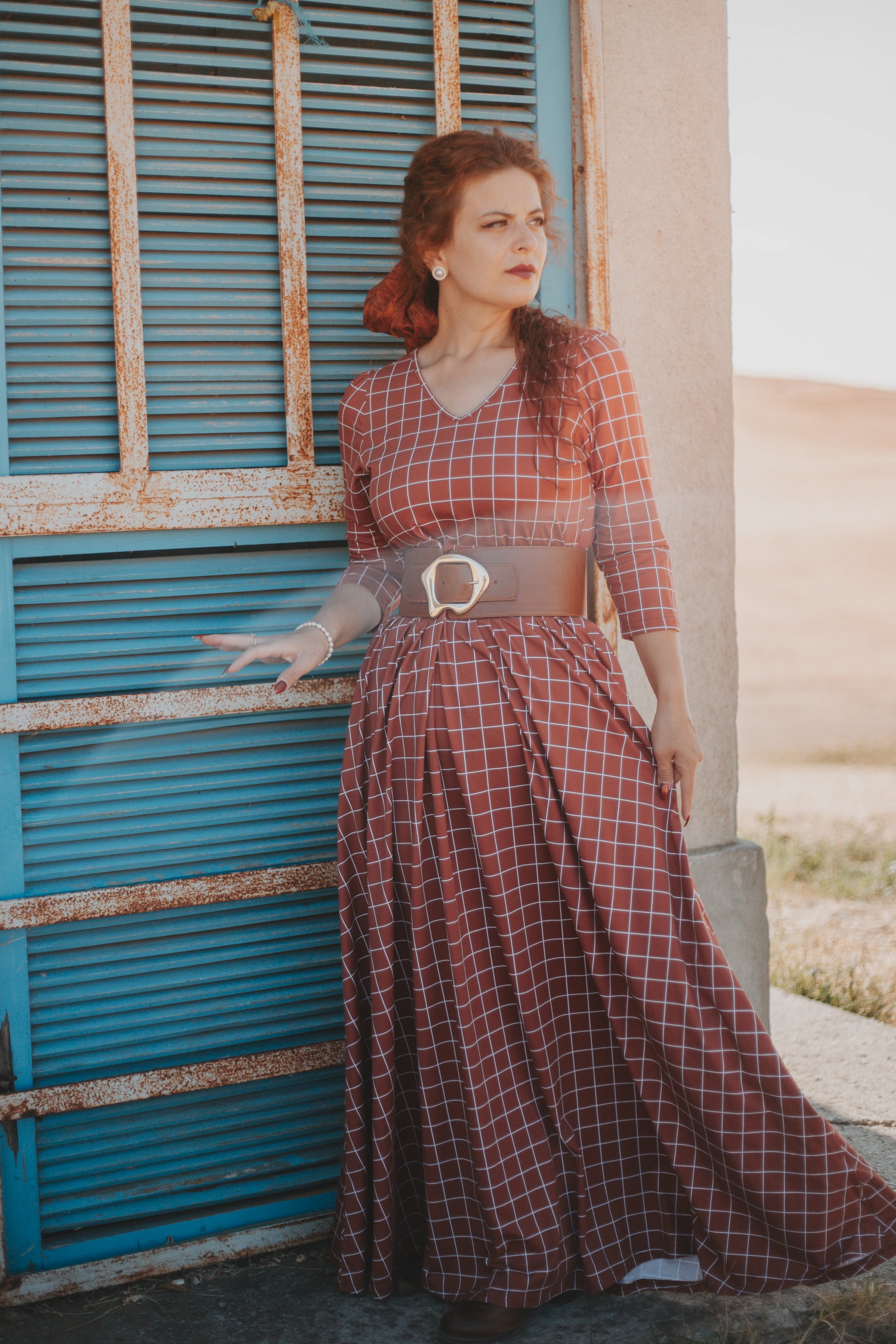 Woman wearing modest copper windowpane plaid maxi dress with 3/4 sleeves and v-neckline, standing elegantly by rustic blue shuttered door, demonstrating the nursing-friendly design and versatile style perfect for casual and formal occasions