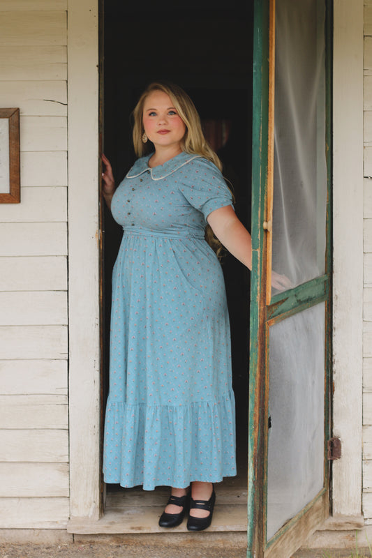 Woman in a blue modest nursing dress standing in a doorway of a rustic building.