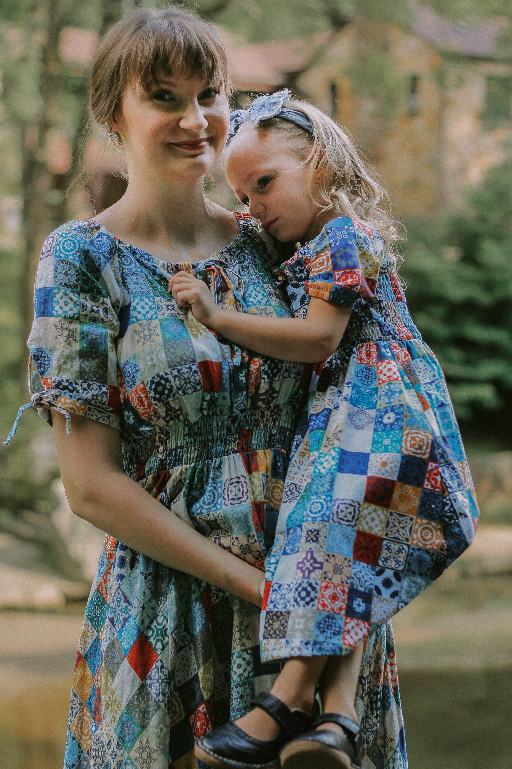 Woman and child wearing colorful patchwork modest dresses outdoors.