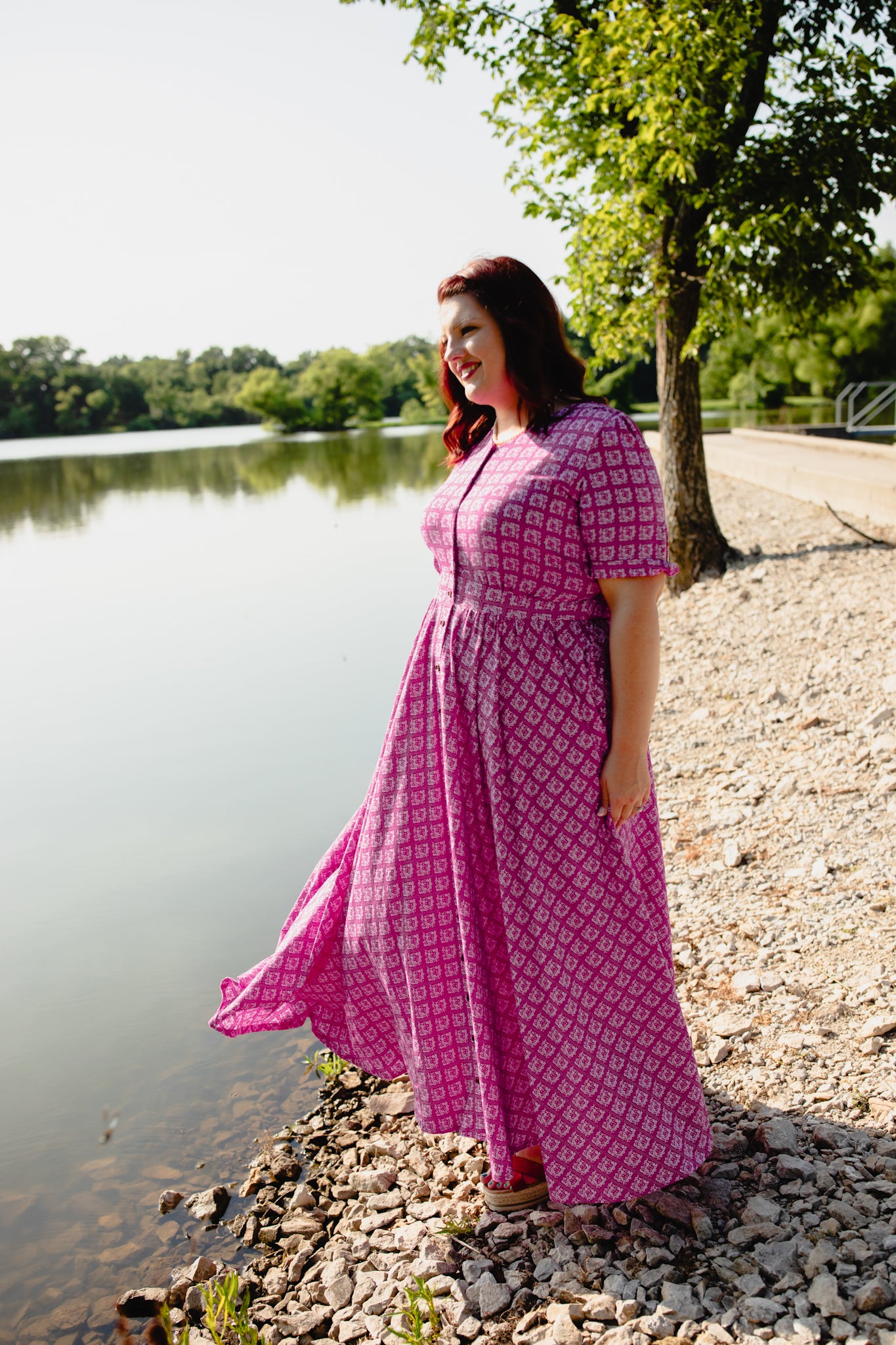 Woman in a pink modest nursing dress standing by a lake with trees in the background