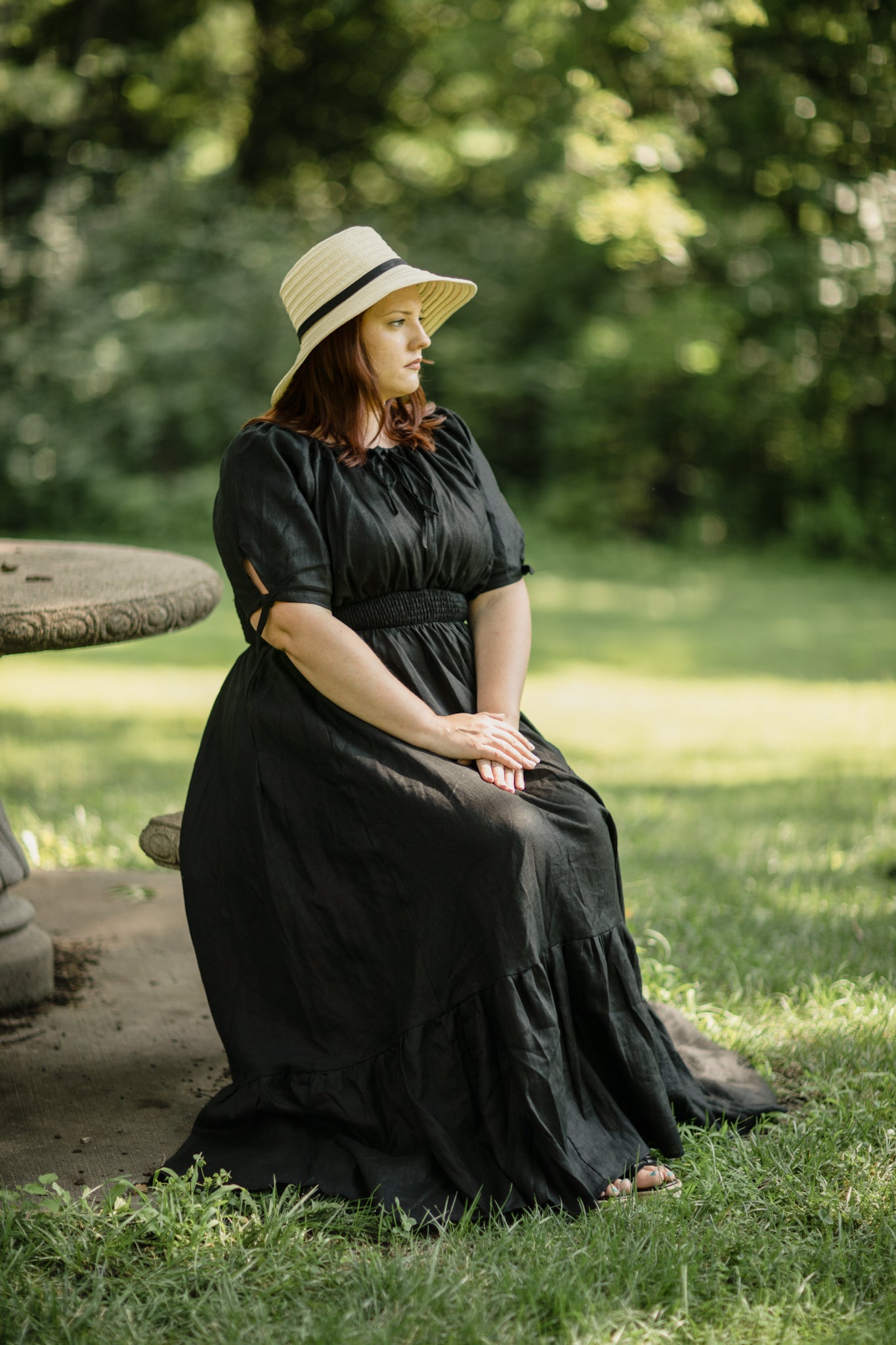 Woman in a black modest nursing dress and straw hat sitting on a stone bench in a park.
