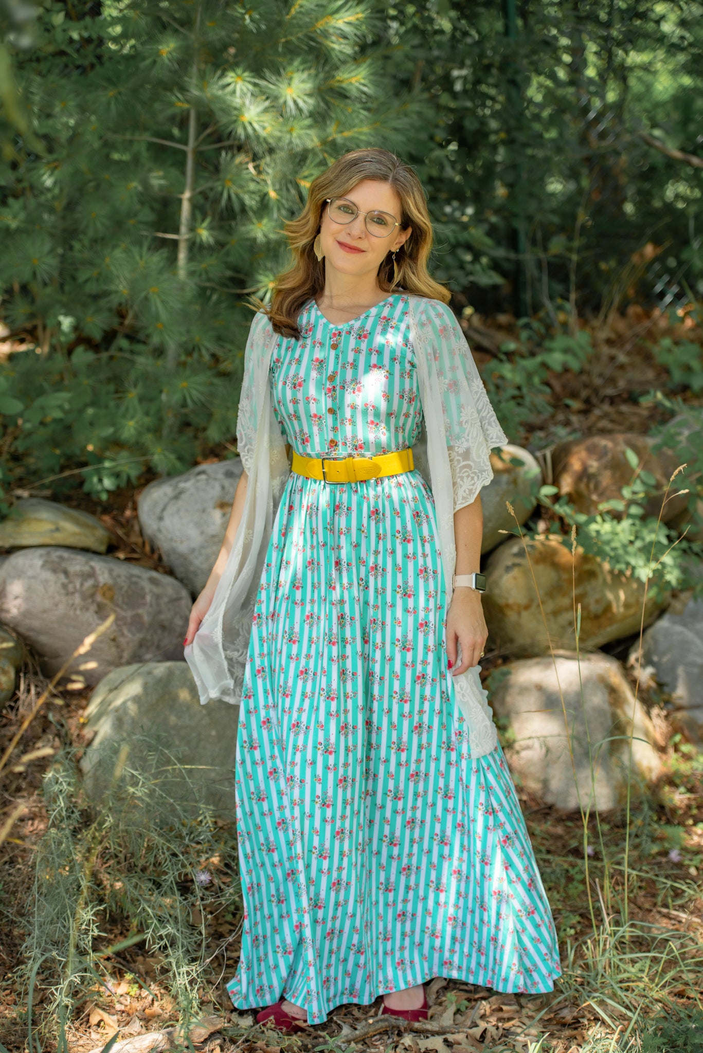 Woman in a turquoise modest dress with a yellow belt standing in a forest setting.