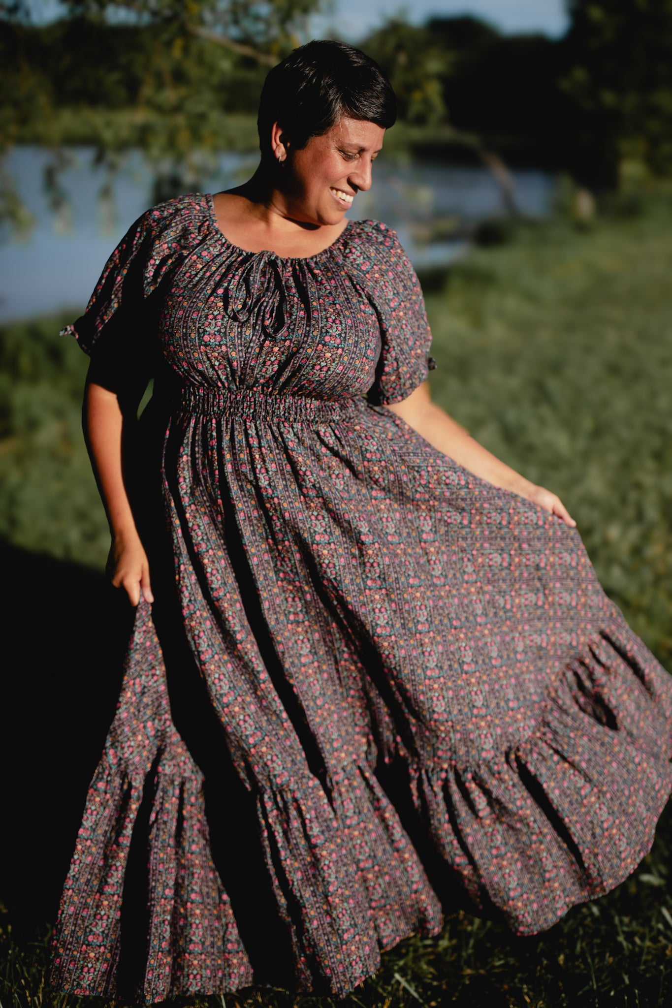 Woman in a long, patterned modest nursing dress standing outdoors with greenery and water in the background
