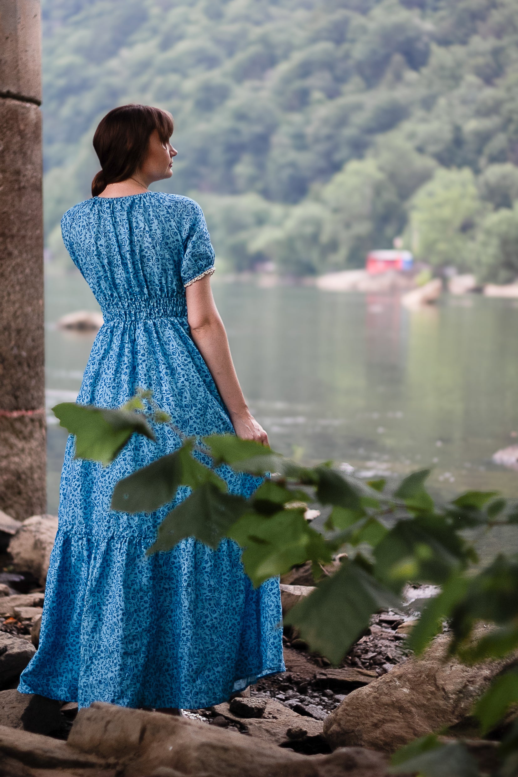 Woman in a blue modest nursing dress standing by a lake with mountains in the background