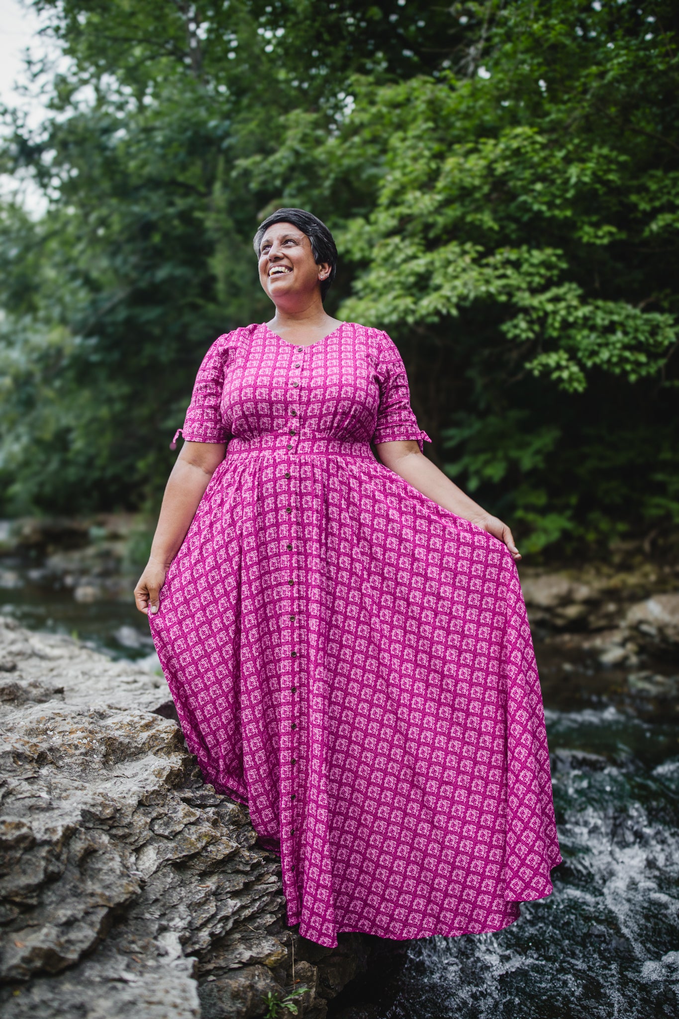 Woman in a pink modest nursing dress standing by a stream with trees in the background