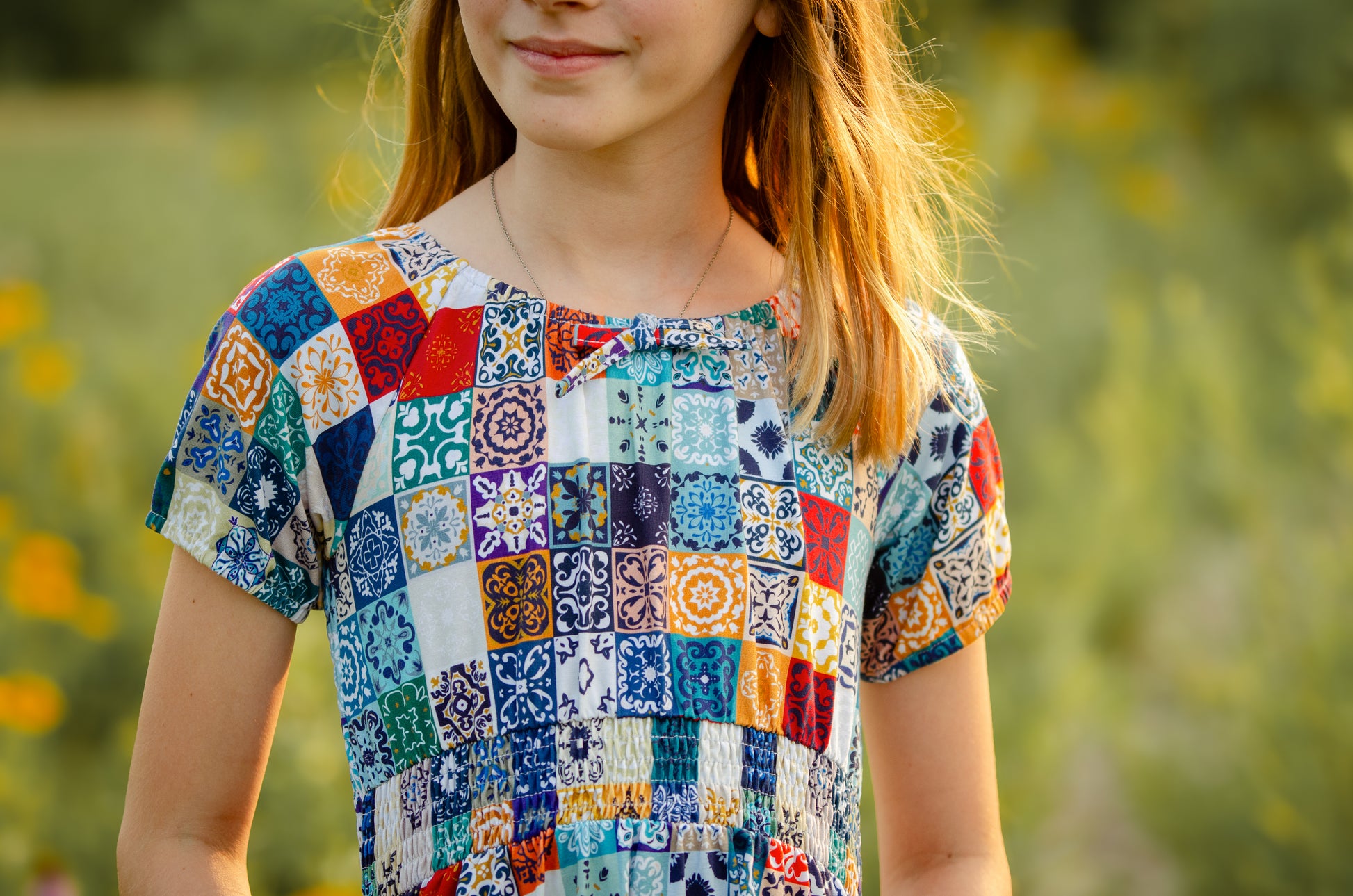 Girl wearing a colorful patchwork modest dress standing in a field with blurred greenery in the background