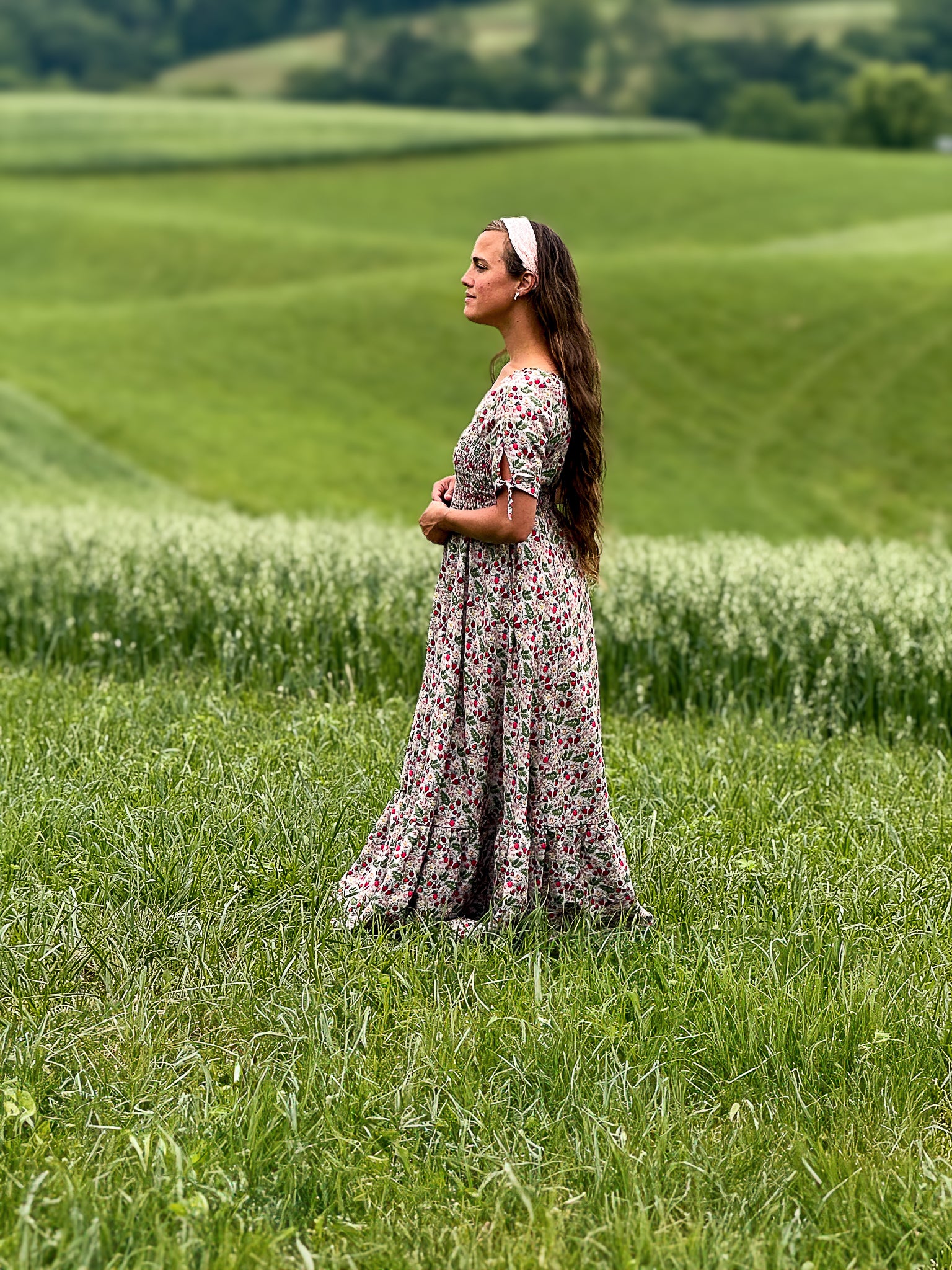 Woman in a floral modest nursing dress standing in a green field with rolling hills in the background