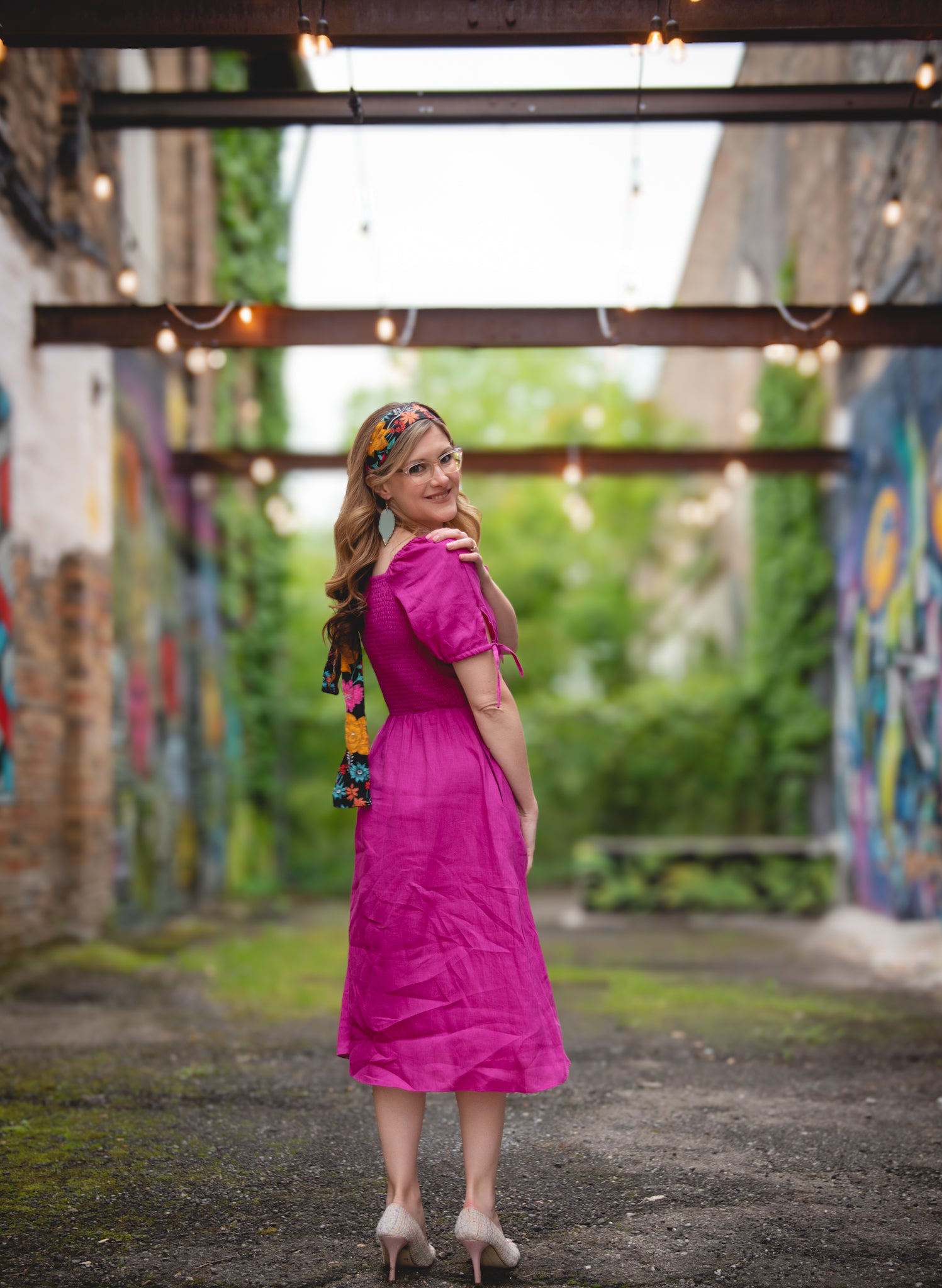 Woman in a pink modest nursing dress standing in an outdoor setting with colorful murals and hanging lights.
