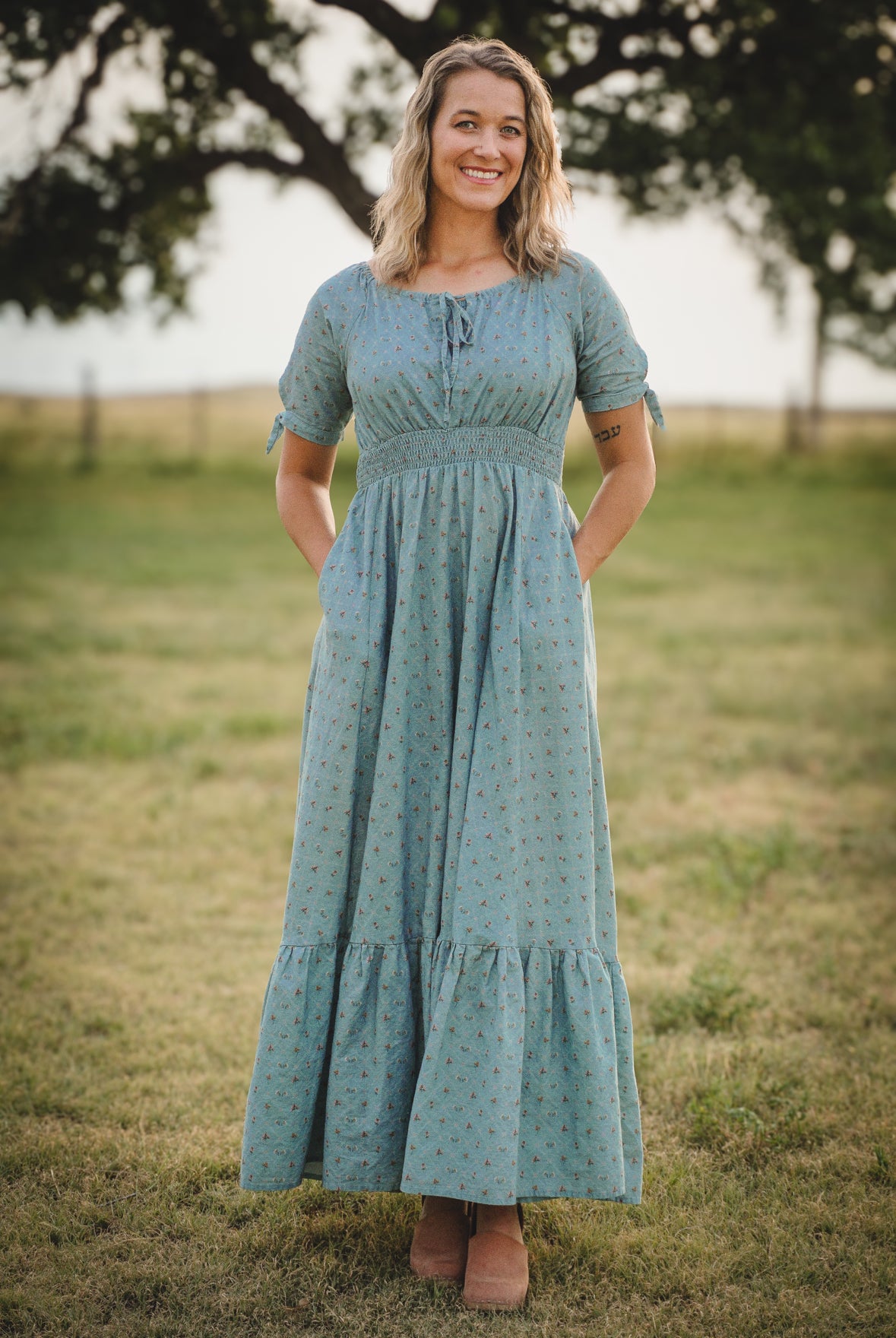 Woman wearing a blue modest nursing dress standing in a grassy field with trees in the background