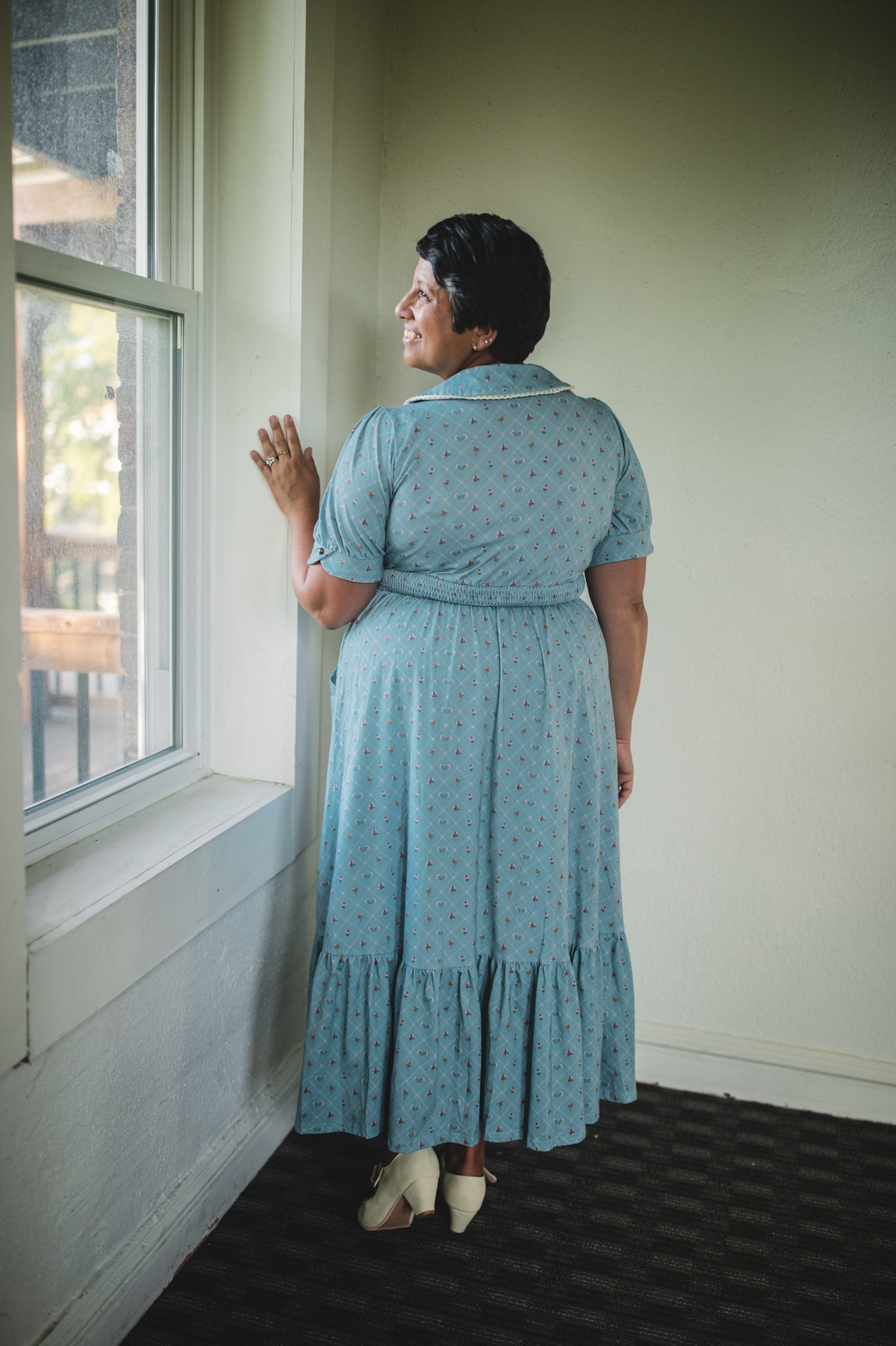 Woman in a modest nursing dress standing by a window, looking out.